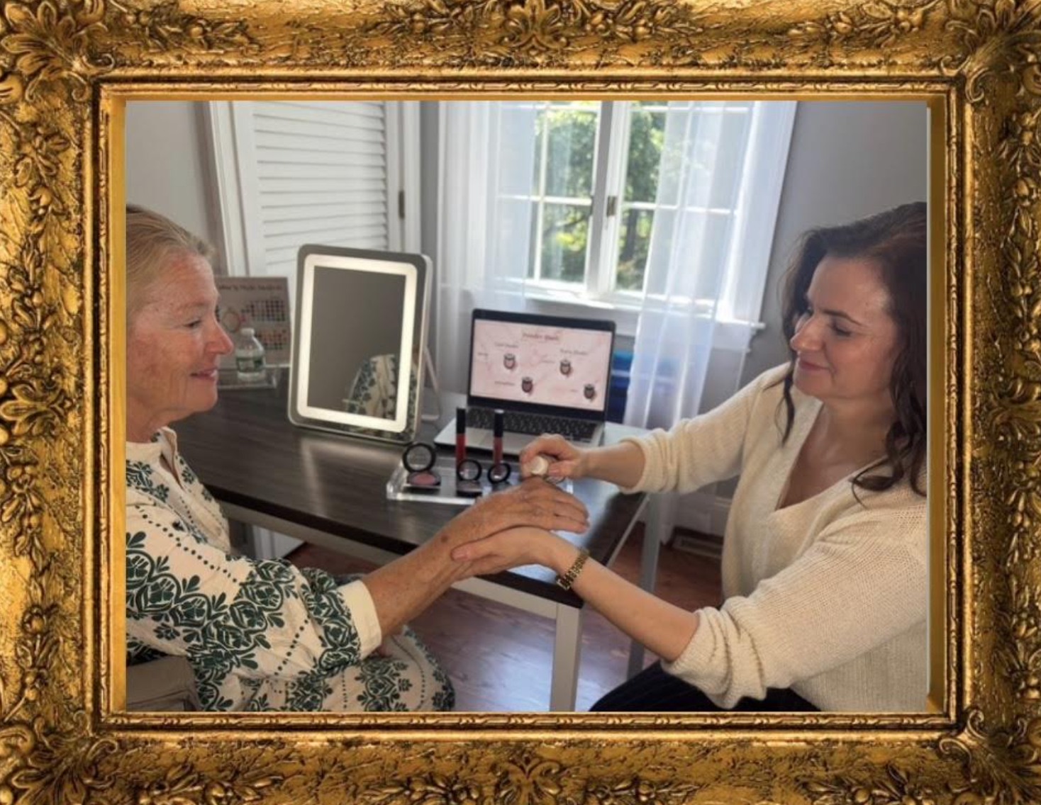 A young woman providing a beauty treatment or skincare consultation to an elderly woman in a well-lit room with windows. The young woman is holding the elderly woman's hand and appears to be applying a product or conducting an examination, with beauty products and a computer screen on the desk.