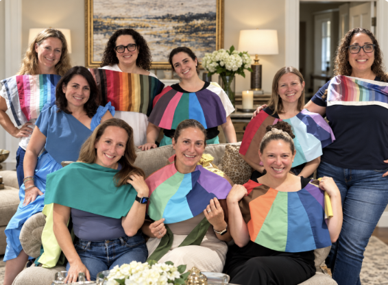 A group of women, some wearing colorful, rainbow-themed fabric pieces on their shoulders or headbands, gathered together indoors for a photo, smiling happily.