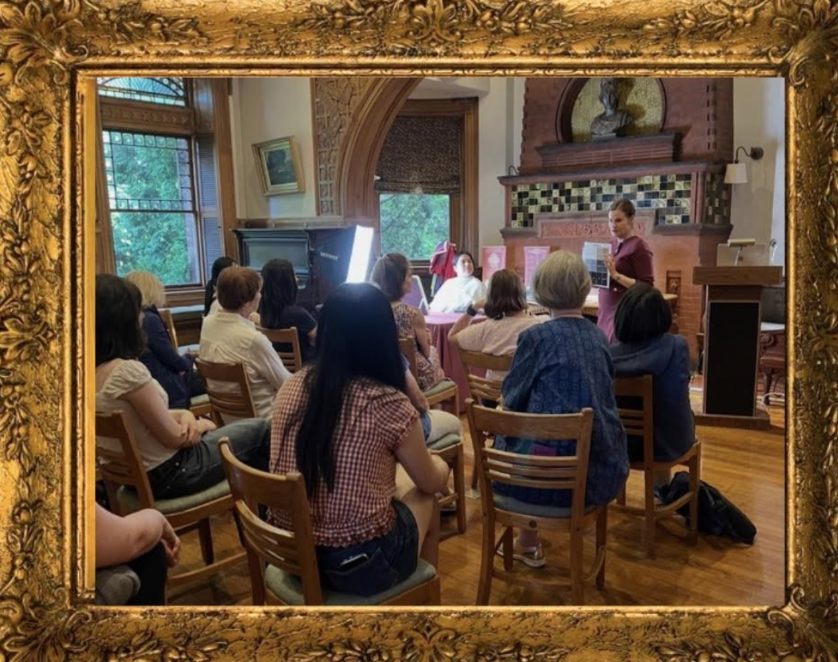 A woman giving a presentation to a seated audience in a decorated room with large windows and wooden flooring.