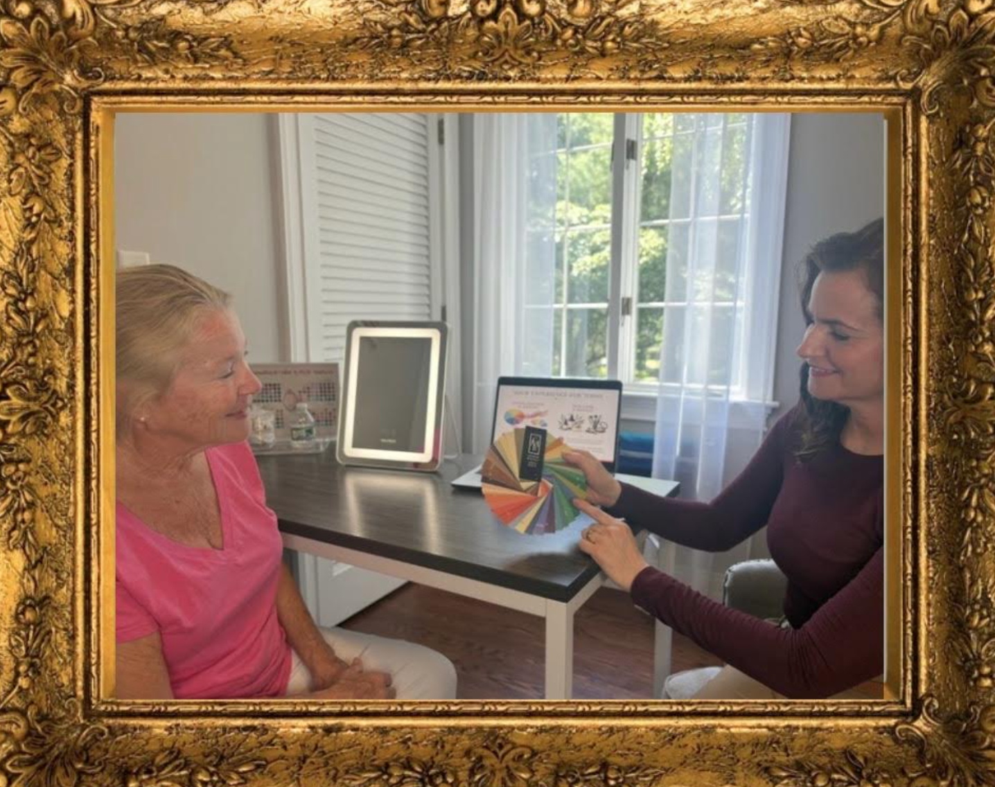 Two women sitting at a table, with the younger woman showing the older woman a palette of various paint colors. There is a window with white curtains behind them, and a digital tablet on the table. The scene is framed with an ornate gold picture frame.