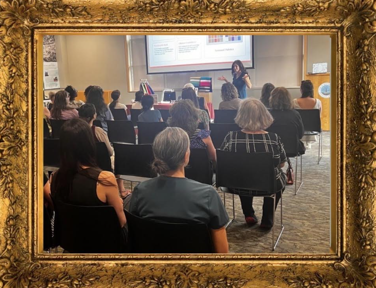 A woman giving a presentation in a conference room, viewed through a decorative gold picture frame, with an audience of diverse adults seated and listening.
