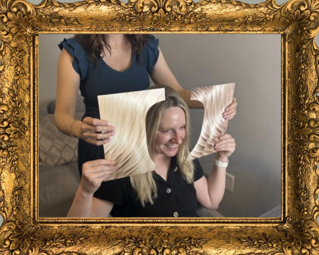 A woman smiling while sitting on a couch, holding a decorative wooden frame over her face with holes for eyes. Another person stands behind her, holding the frame in place.