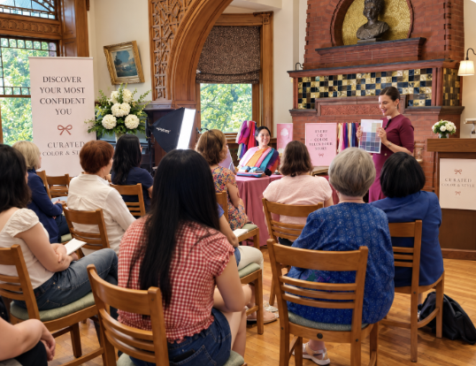 A woman giving a presentation to a seated audience in a decorated room with large windows and wooden flooring.