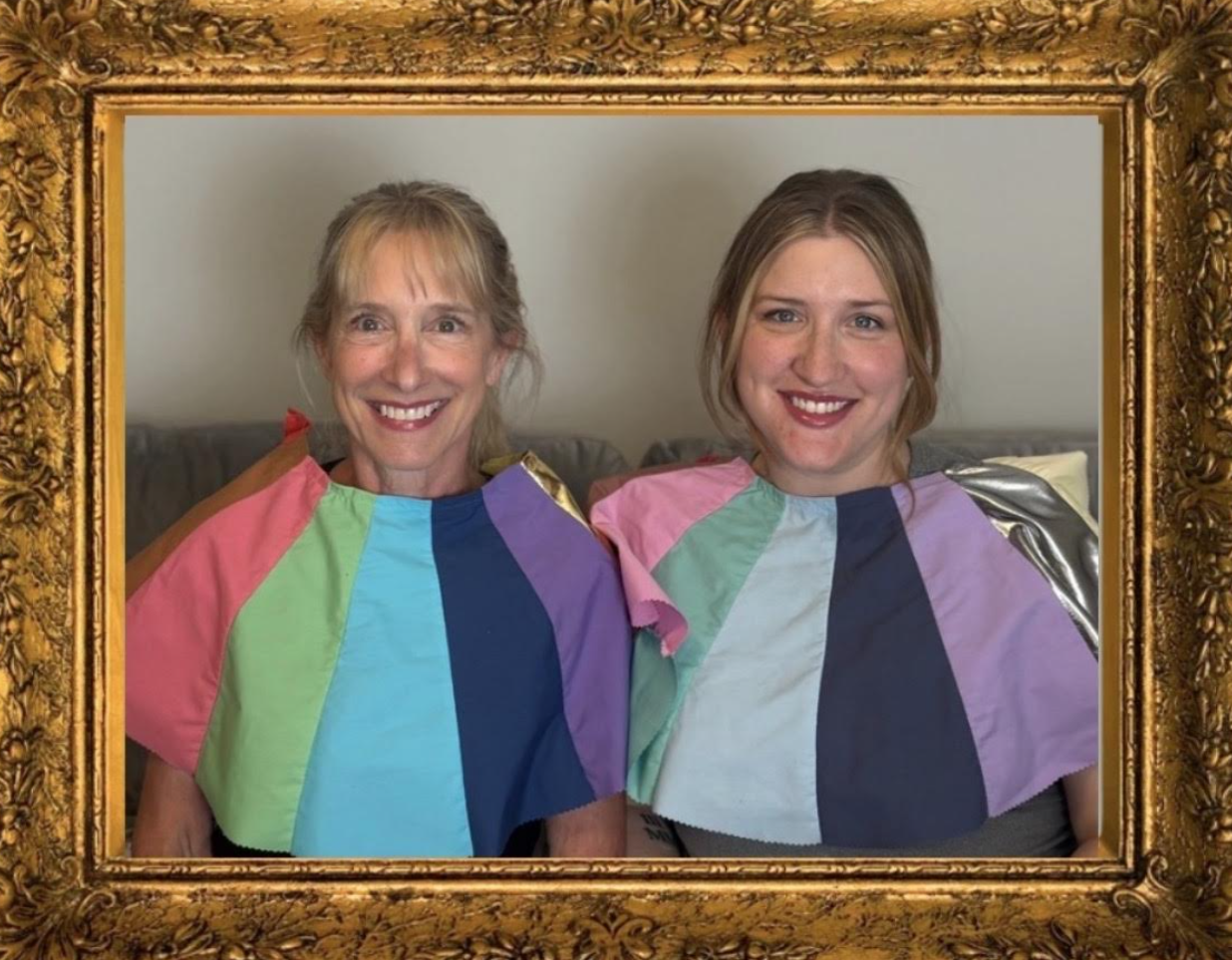Two women smiling, sitting on a couch, with jewel-tone rainbow-colored capes over their shoulders, framed in an ornate gold picture frame.