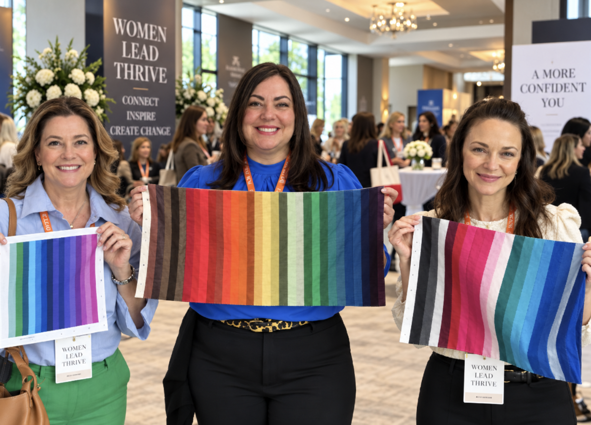 Three women standing together at an event, holding rainbow and color-striped fabric samples, framed by an ornate gold frame.