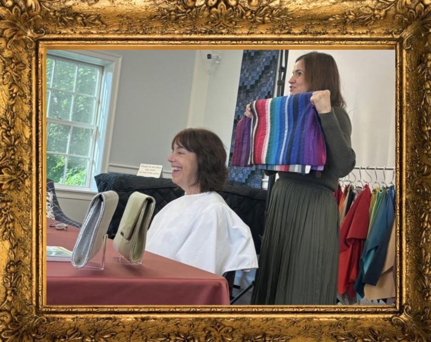 A woman with brown hair holding a colorful striped fabric, smiling at another woman with brown hair in a white apron, who is sitting at a table with fabric samples.