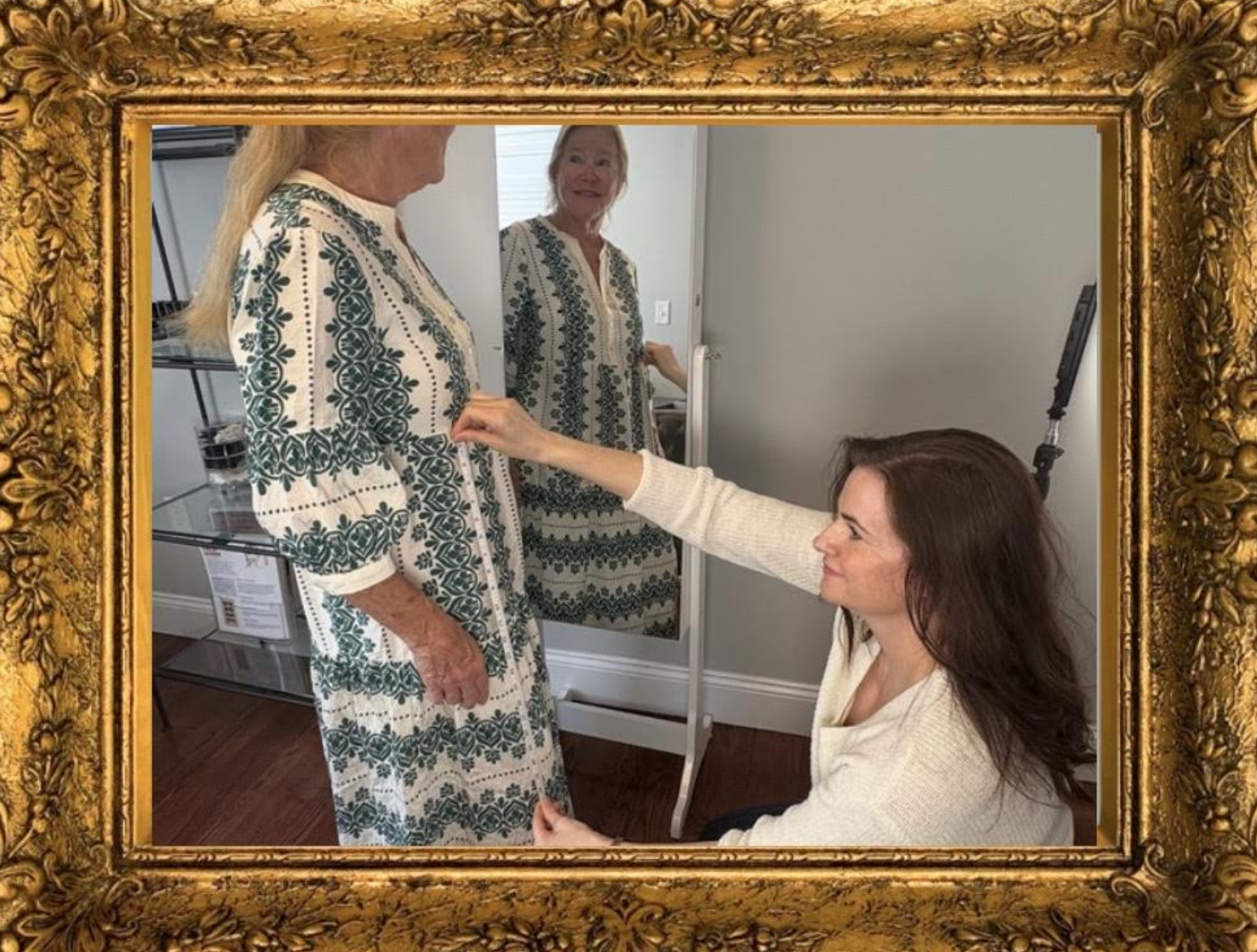 A woman kneels in front of an elderly woman, adjusting her dress while smiling. Both women are wearing patterned dresses and are in a room with a mirror and a glass display shelf.