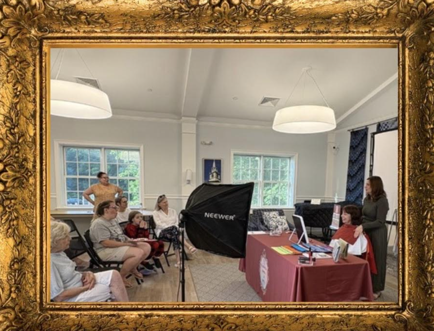 A group of people attending a presentation or workshop in a light-colored room with large windows, framed by an ornate gold picture frame. One woman stands at a table with a computer, while others are seated and listening. A large softbox light is set up for photography or video.