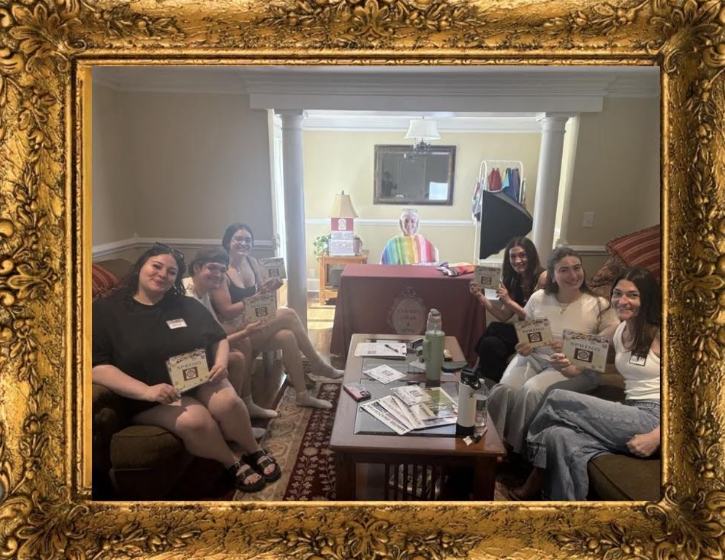 Six women sitting on couches inside a living room, holding flyers, with a woman in rainbow-striped clothing seated at a table in the background. The room has beige walls, white trim, and a large mirror on the wall.