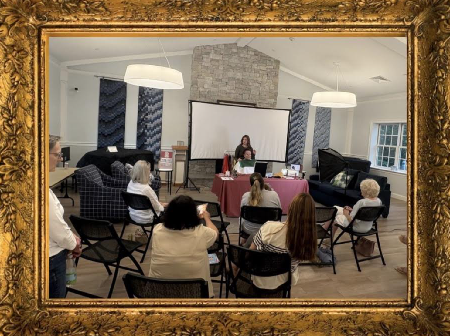 A group of people in a room with a presentation setup, seated on black chairs, facing a woman standing in front of a projection screen. The room has a stone fireplace, a black couch, large windows, and hanging lights. The photo is framed with an ornate gold frame.