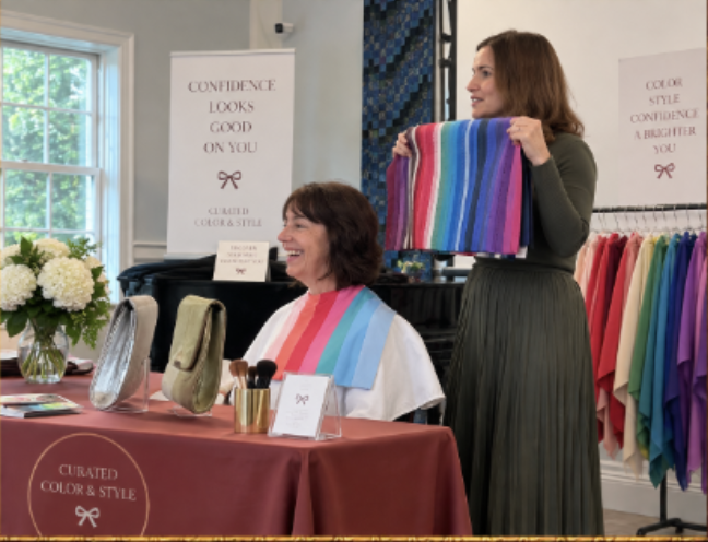 A woman with brown hair holding a colorful striped fabric, smiling at another woman with brown hair in a white apron, who is sitting at a table with fabric samples.