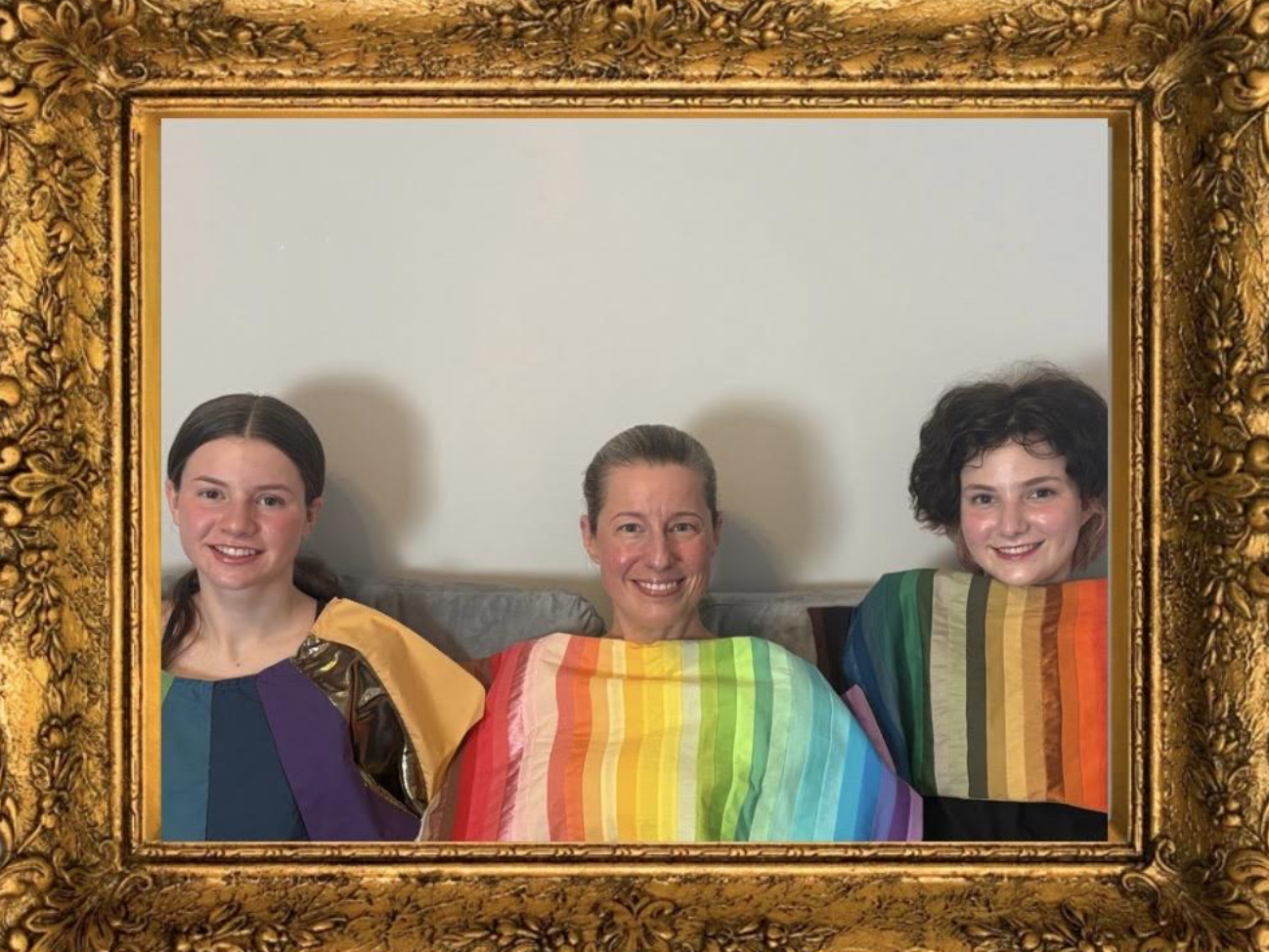 Three women sitting on a couch, smiling, viewed through an ornate gold picture frame. They are wearing rainbow-colored garments.