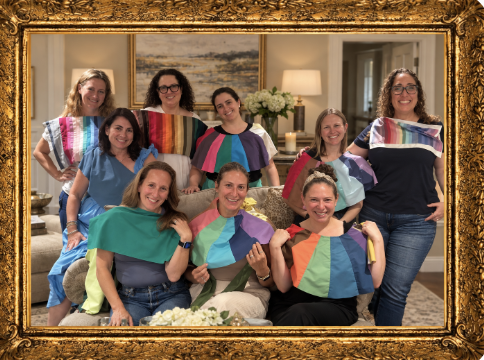 A group of women, some wearing colorful, rainbow-themed fabric pieces on their shoulders or headbands, gathered together indoors for a photo, smiling happily.
