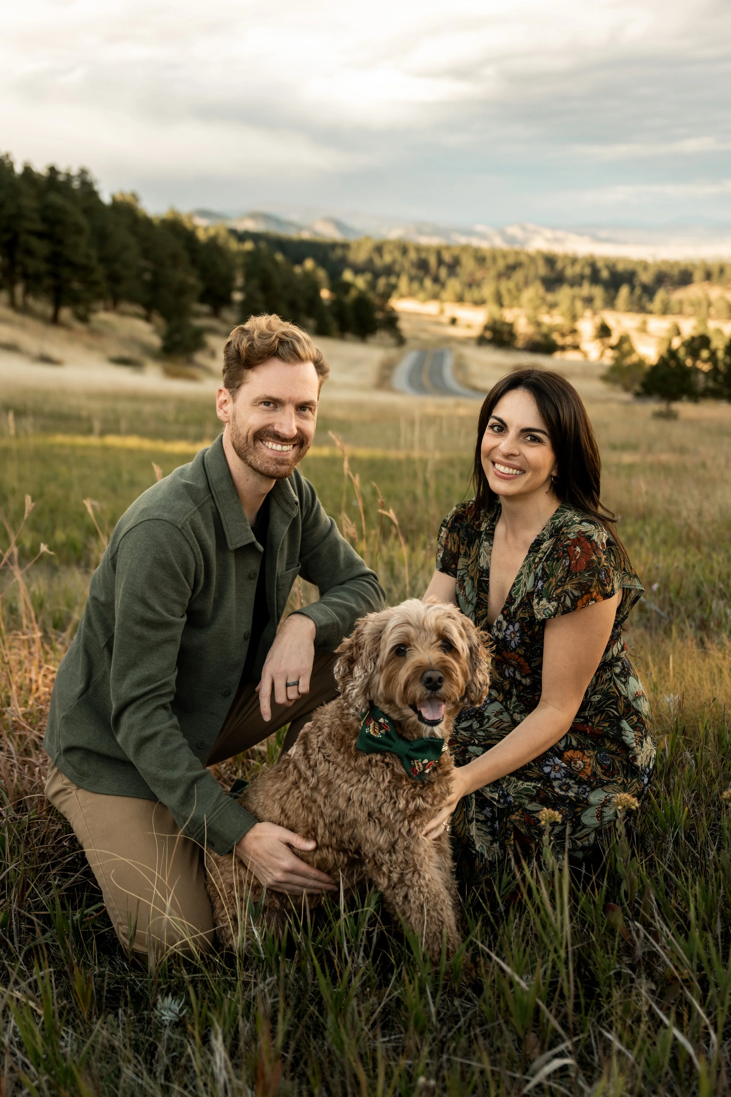 A smiling couple with a fluffy brown dog sitting in a grassy field with trees and mountains in the background.
