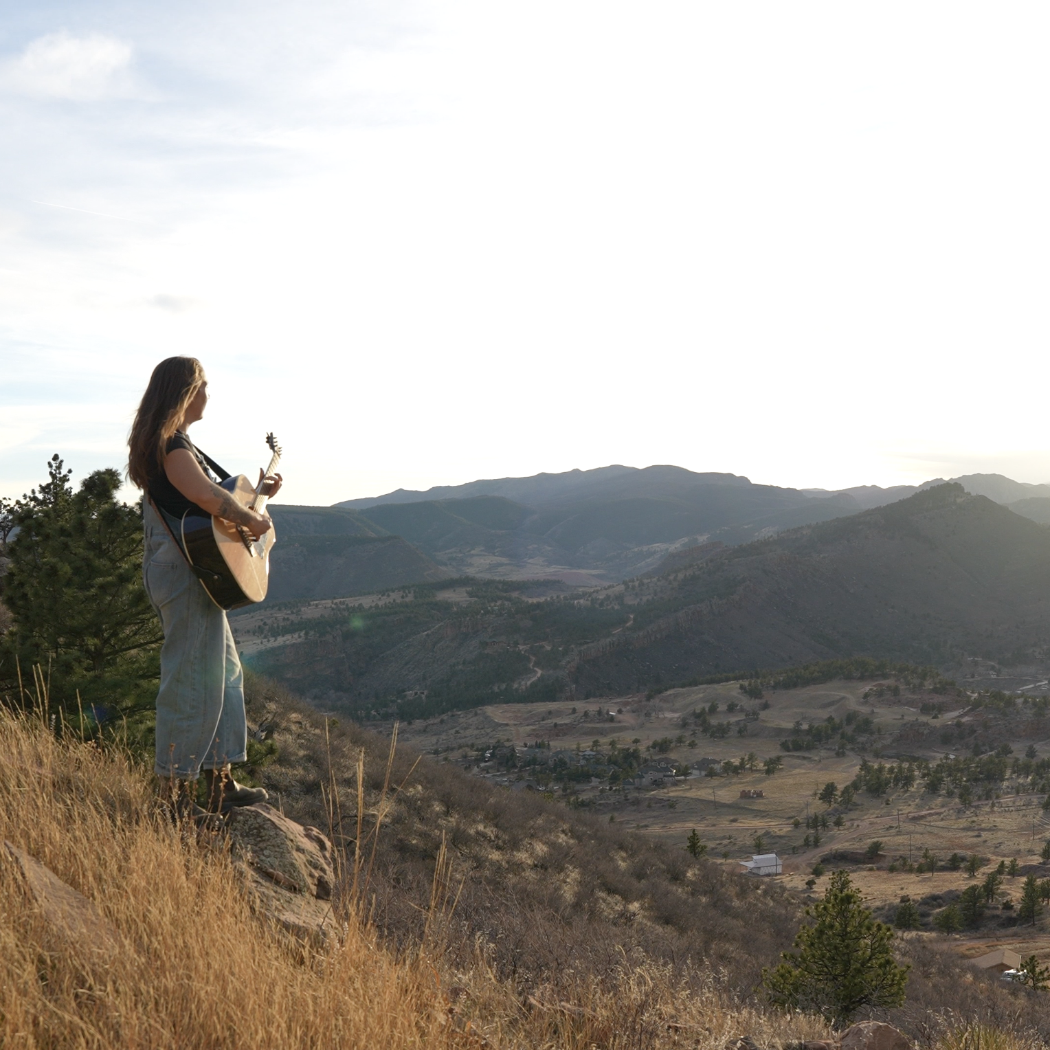 A woman stands on a hill playing an acoustic guitar, overlooking a valley with mountains in the background during sunset.