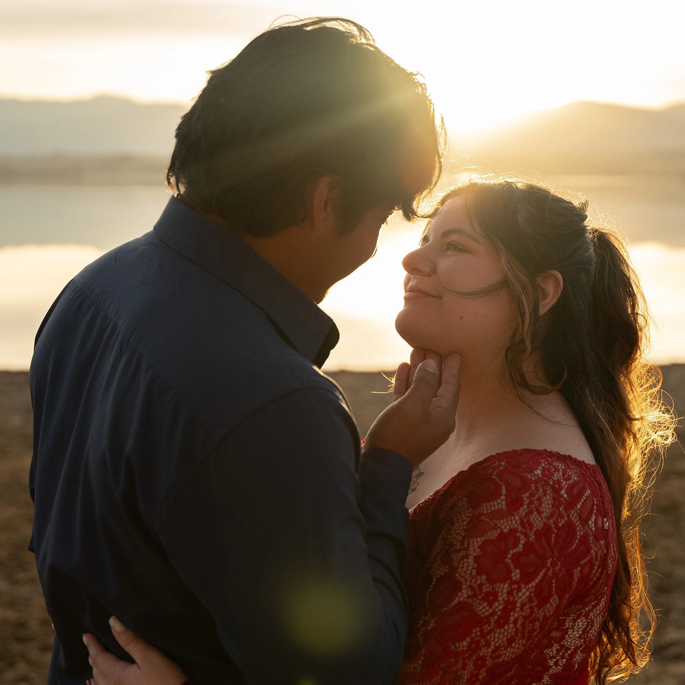 A man and a woman sharing an intimate moment on the beach at sunset, gazing into each other's eyes with the man holding the woman's chin gently.
