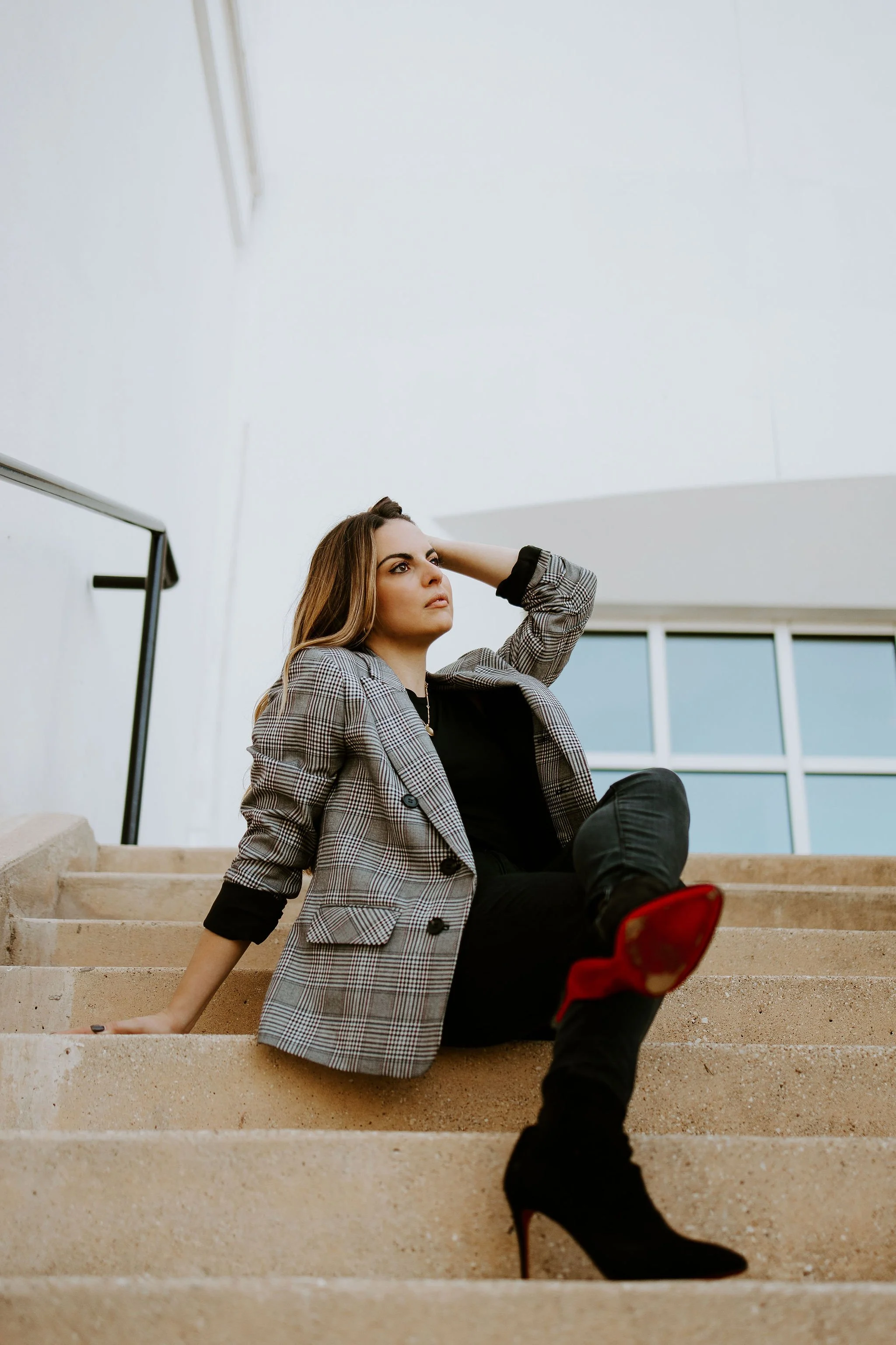 A woman with brown hair sitting on beige stairs indoors, wearing a checkered blazer, black pants, and black high-heeled boots with red soles, looking thoughtful with her hand resting behind her head.