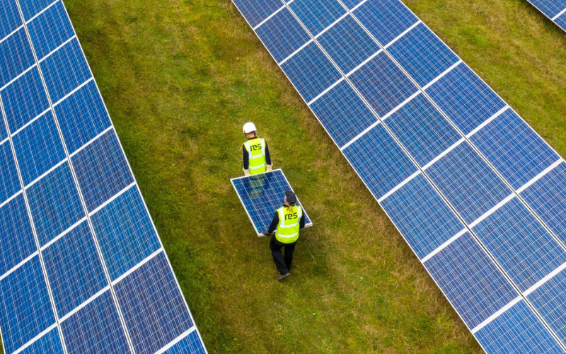 Two workers in safety vests and helmets installing or inspecting solar panels in a solar farm with rows of blue solar panels on grassy ground.