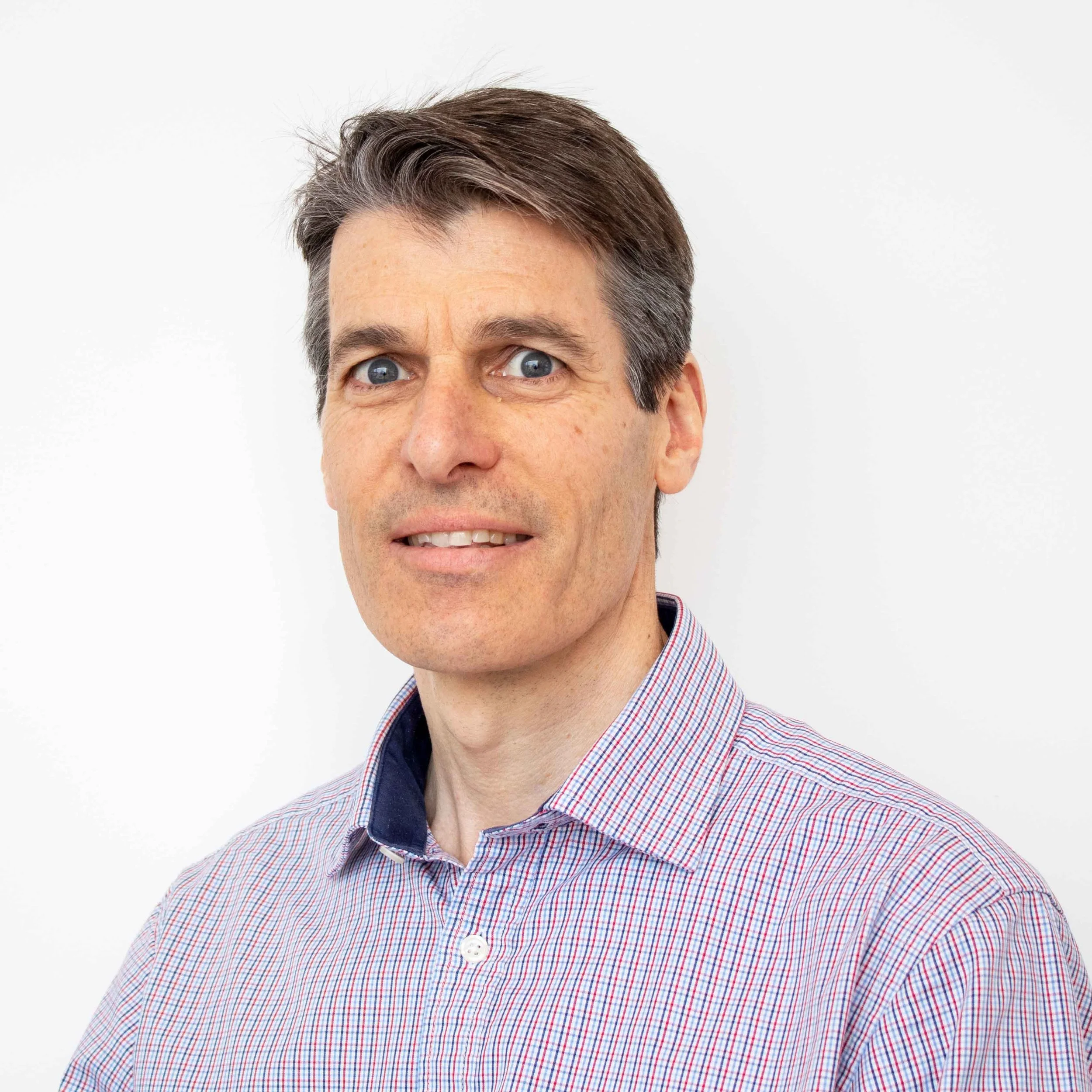 A middle-aged man with short brown hair, blue eyes, and light skin, wearing a checkered button-down shirt, looking at the camera against a plain white background.