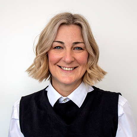 A smiling woman with short blonde hair, wearing a white shirt and a black sleeveless sweater, standing against a plain white background.