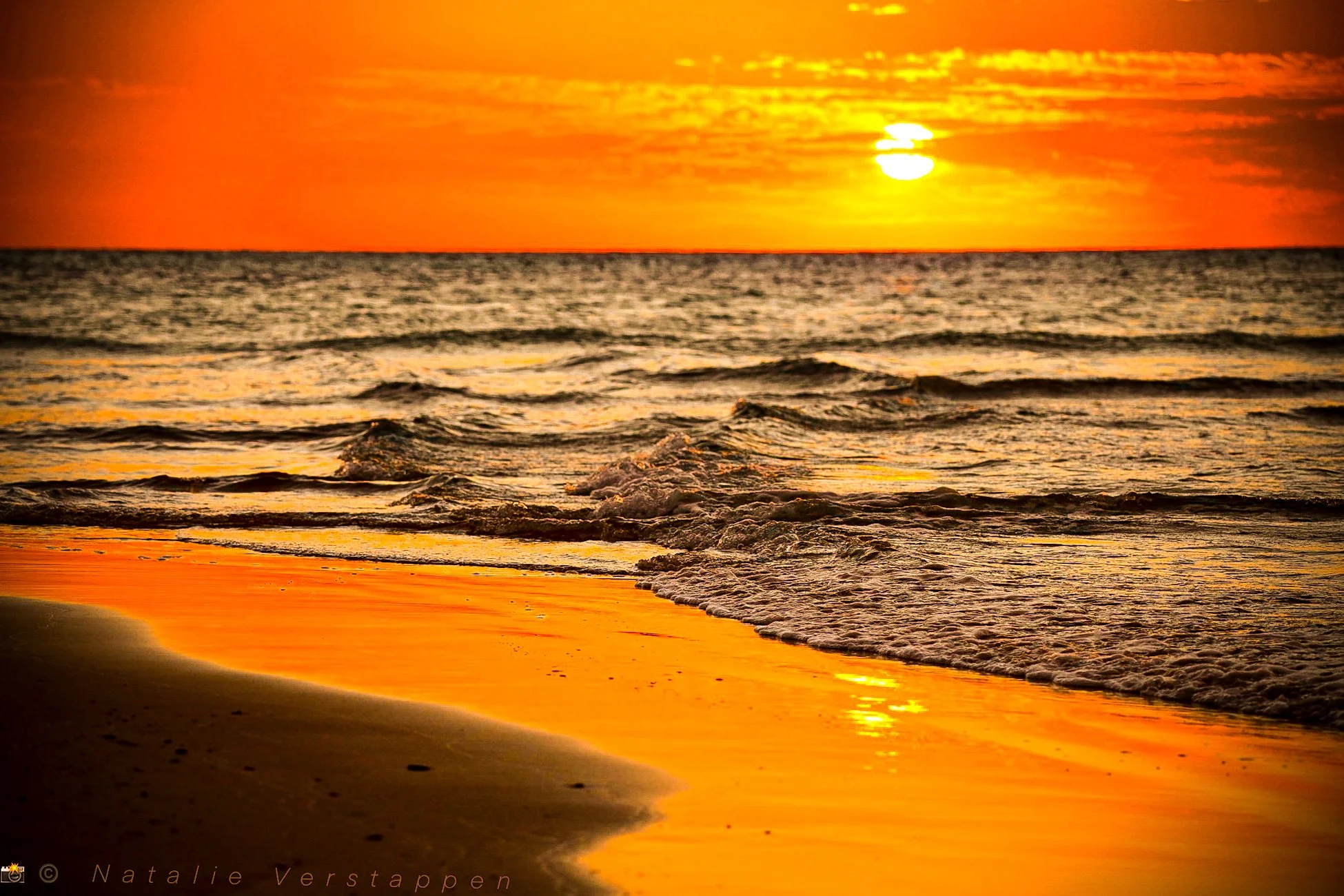 A sunset over the ocean with an orange and yellow sky, and waves gently crashing on the sandy beach, reflecting the warm colors.