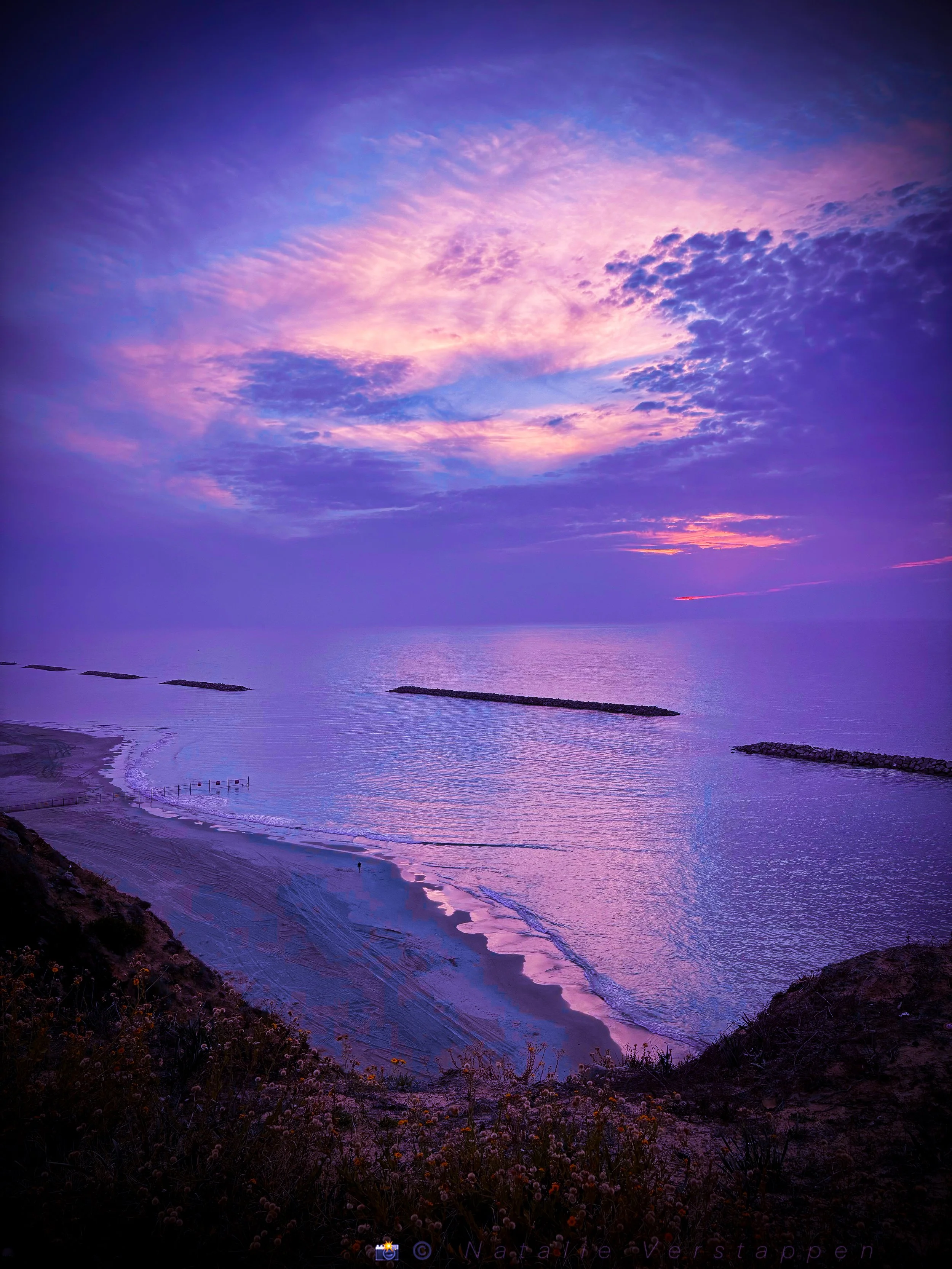 A scenic view of a colorful sunset over the ocean with purple, pink, and blue clouds in the sky and a rocky coastline with patches of vegetation in the foreground.