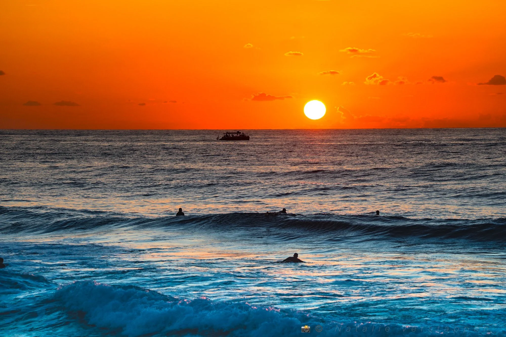 Boat sailing towards the sunset in an ocean filled with surfers