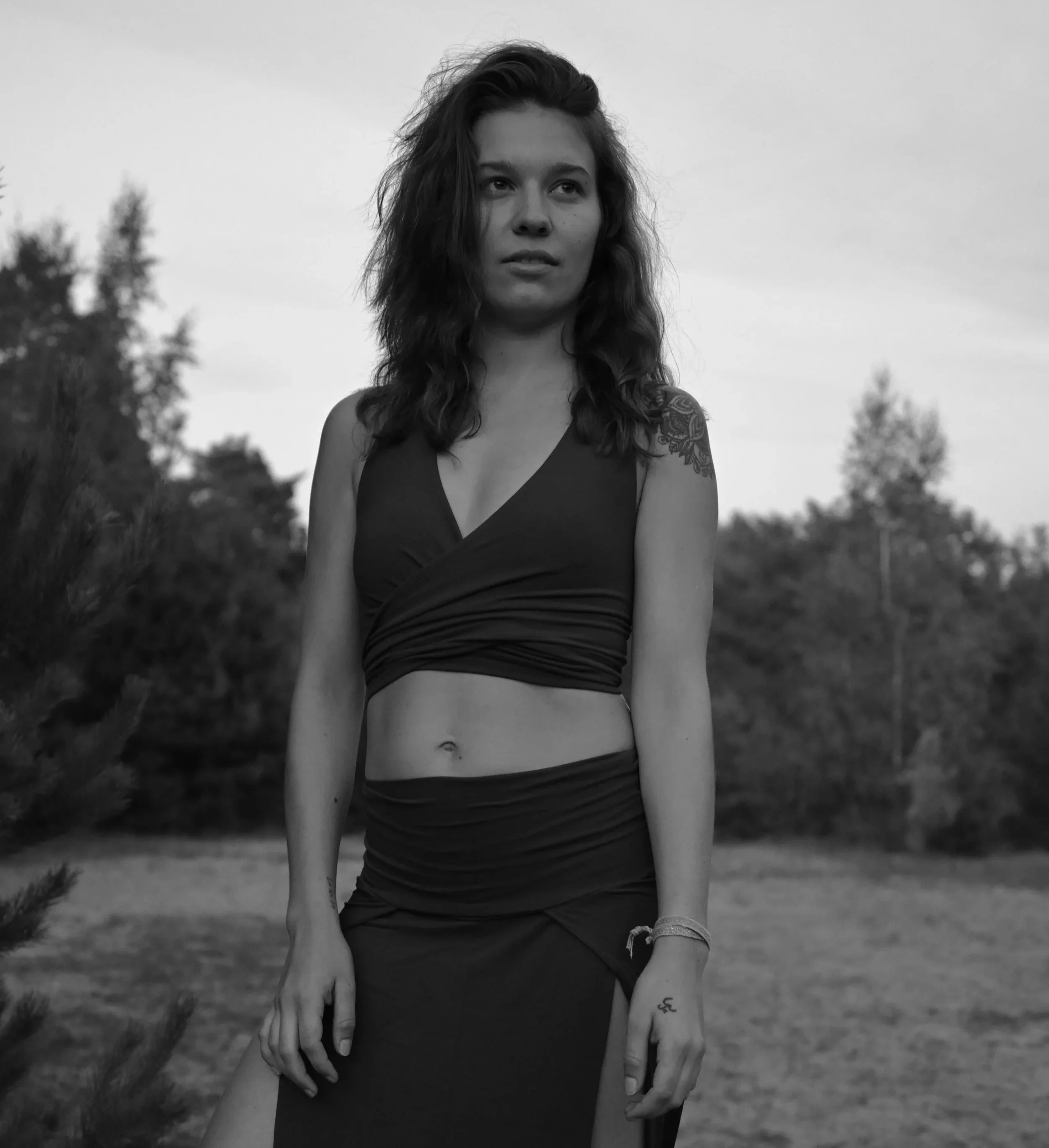 Black and white photo of a young woman outdoors, standing with trees and open sky in the background. She has dark, wavy hair, wears a crop top and a long skirt, and has tattoos on her arm.
