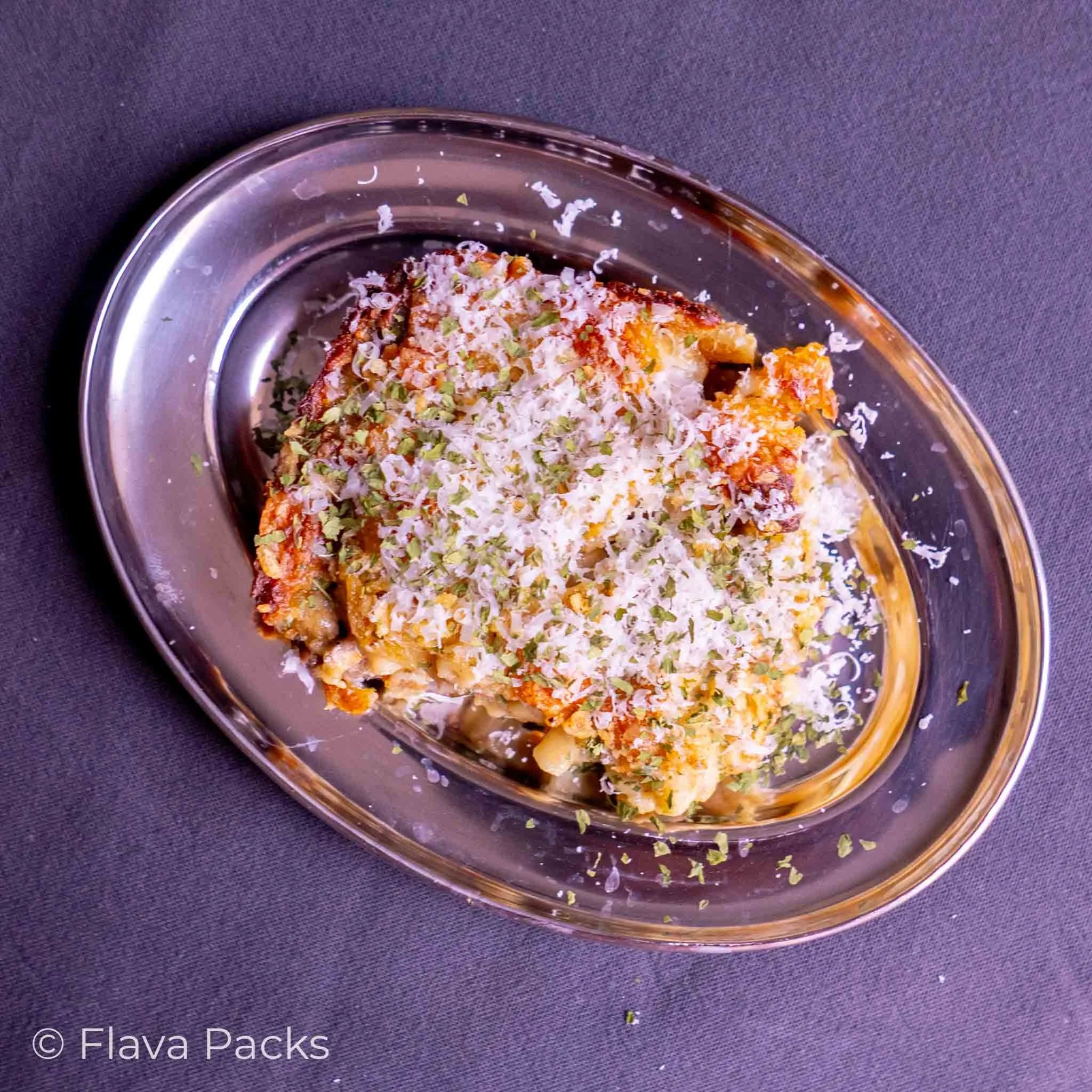 Jerk Lamb Mac & Cheese topped with parmesan on a stainless steel dish with a grey blue background