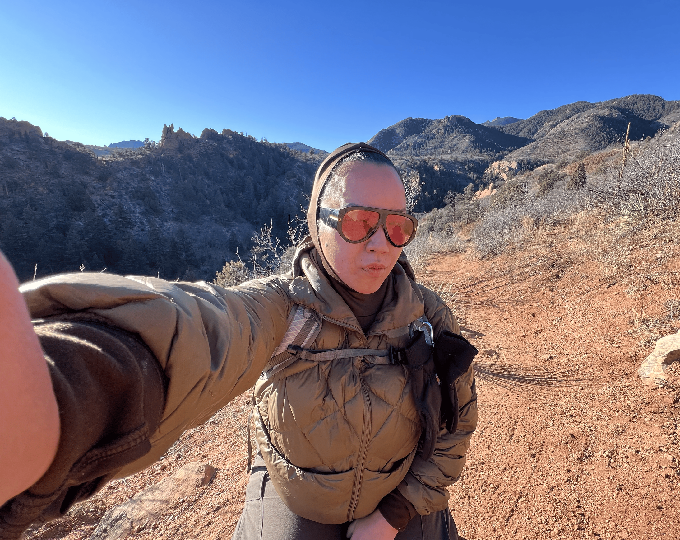 A woman wearing sunglasses, a hoodie, and a jacket taking a selfie in a mountainous outdoor area with clear blue sky and dry vegetation.