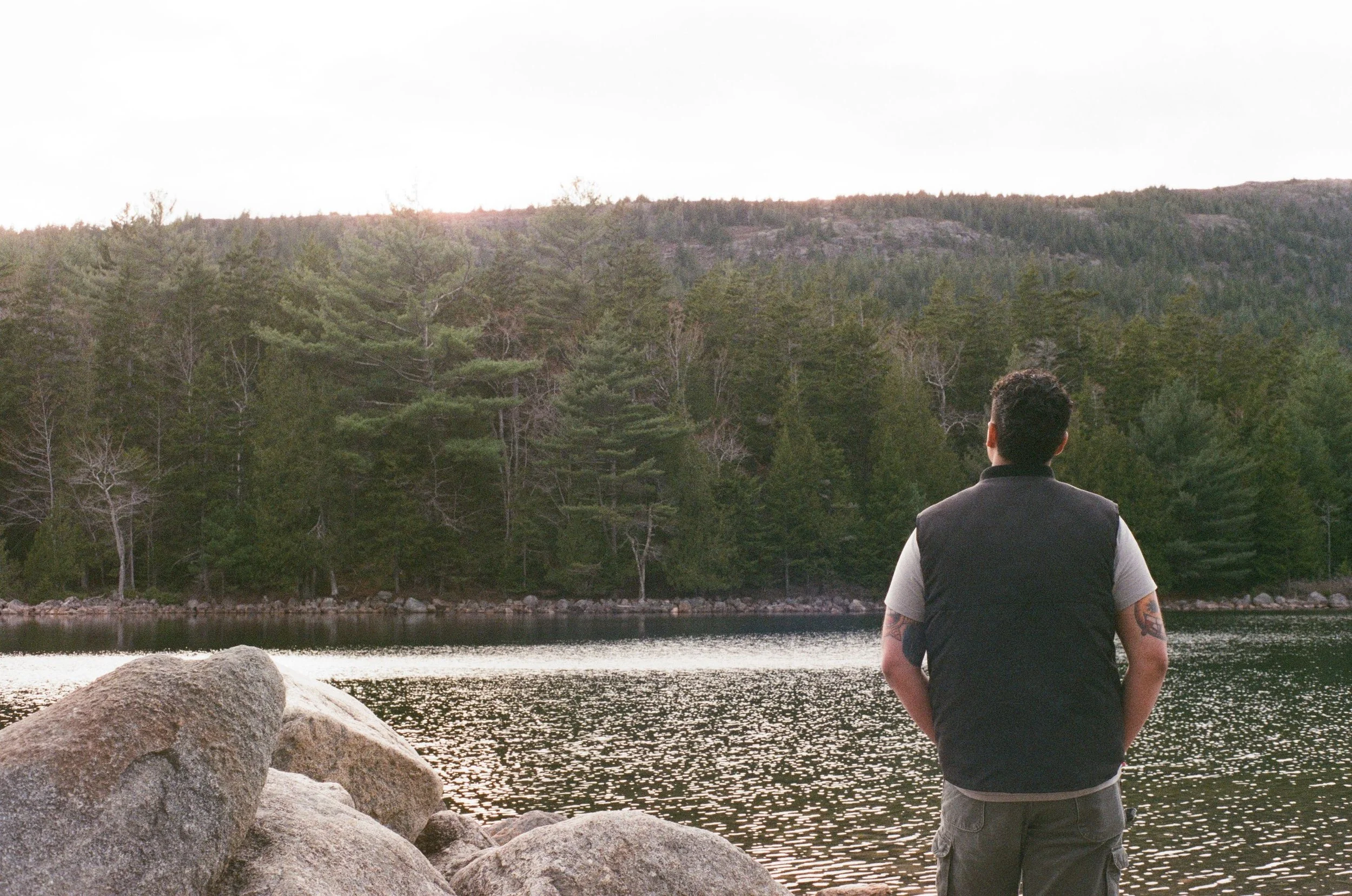 A man with dark hair, tattoos on his arms, wearing a black vest over a gray shirt, stand by a body of water overlooking a dense forest with green trees and rocky hills in the distance during sunset or sunrise.