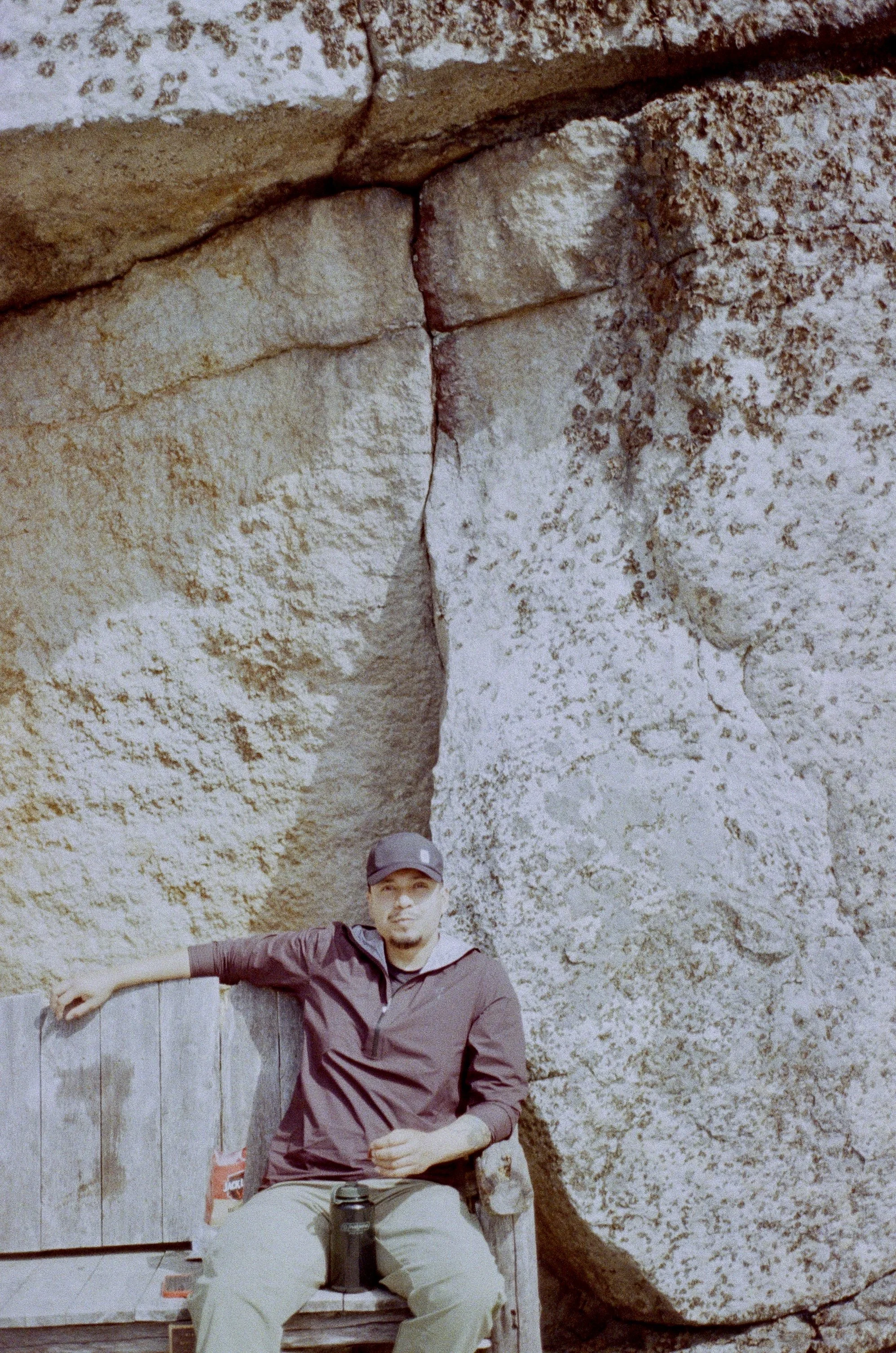 A man sitting on a wooden bench outdoors, wearing a dark jacket, light pants, and a baseball cap, with a large stone rock formation in the background.