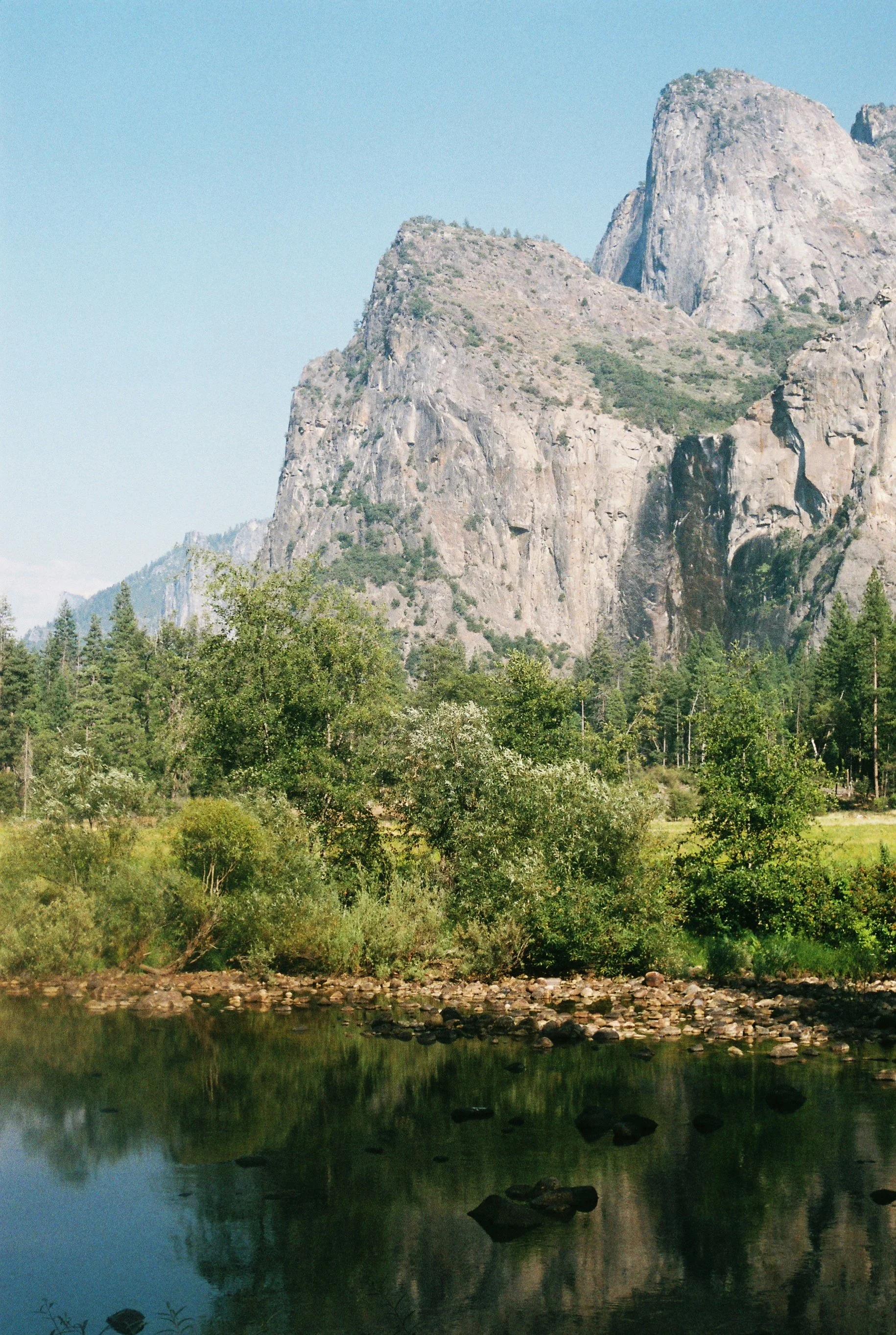 A scenic view of a river or lake with reflections of trees and rocky mountains under a clear blue sky.