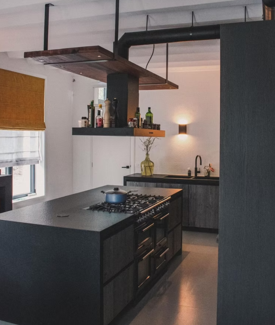 Modern kitchen with black countertops, stainless steel stove, and a black range hood. A window with a yellow and white blind, a small yellow vase with branches, and bottles on a shelf above the stove. There's a black and white sink with a black fauce