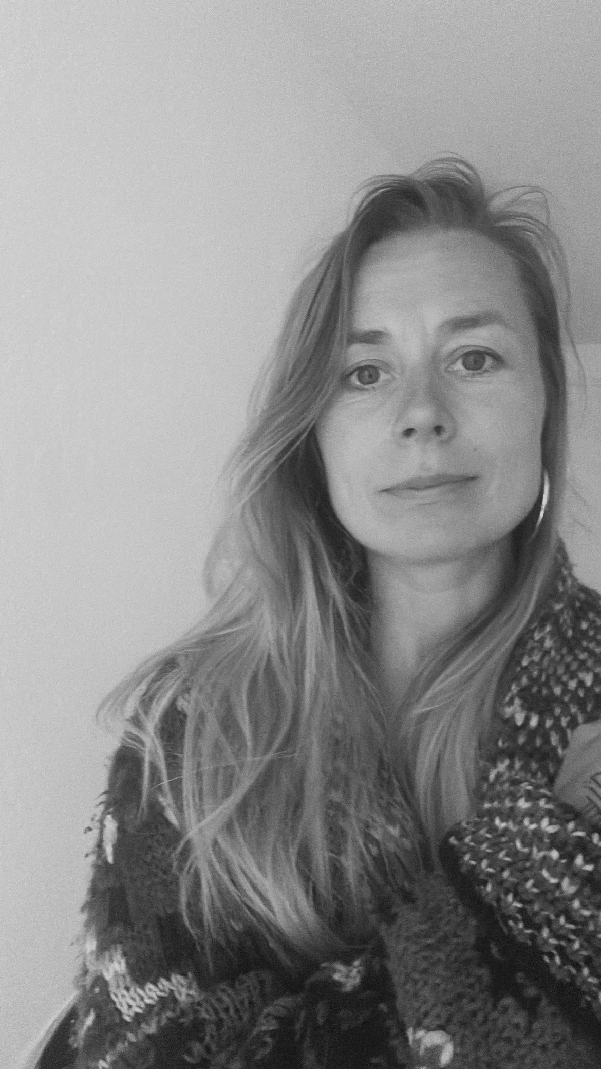 Black and white photograph of a woman with long wavy hair and light eyes, wearing a patterned sweater, looking at the camera.