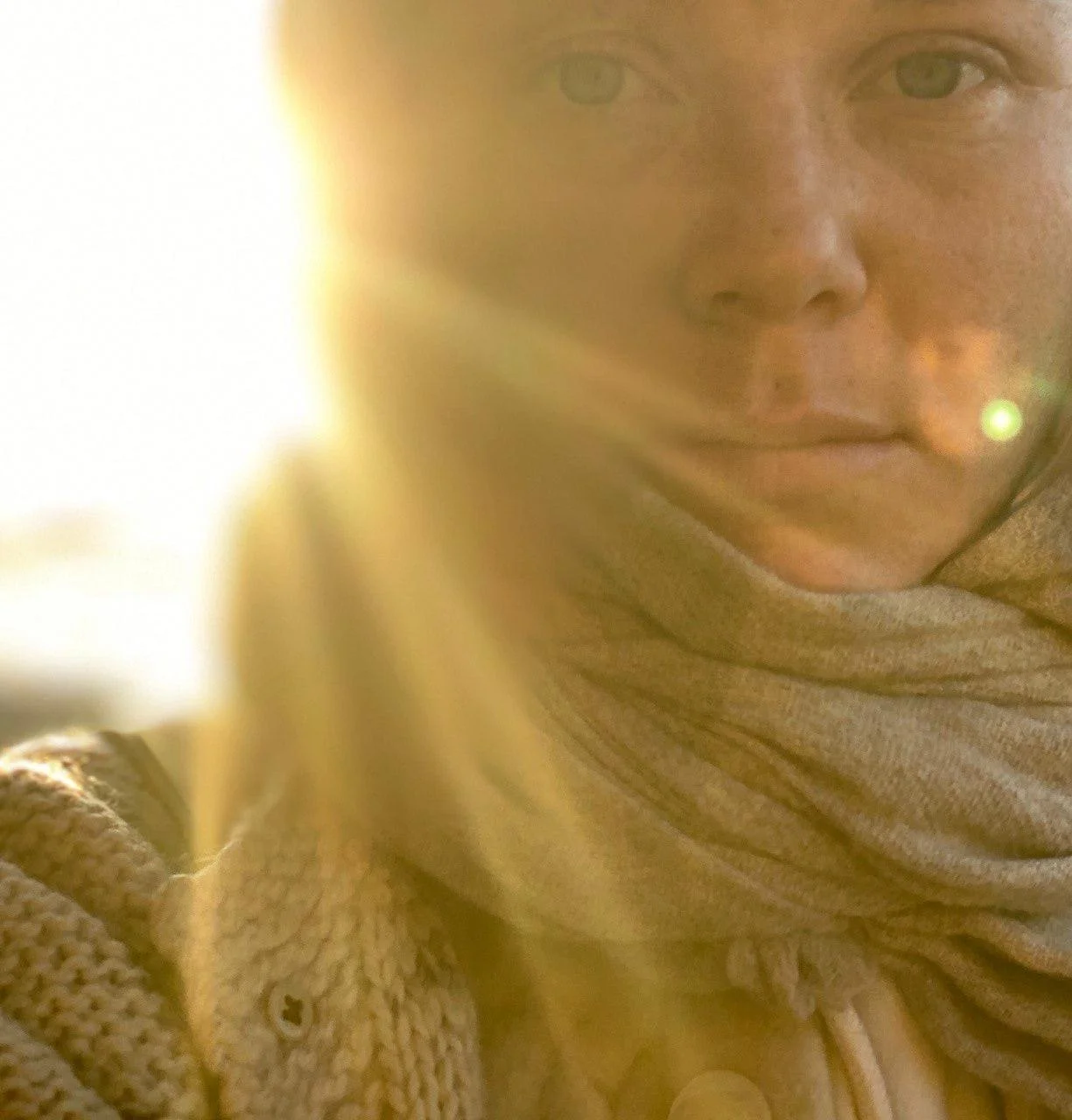 A close-up of a person's face with a neutral expression, wearing a beige scarf, with sunlight creating a lens flare in the image.