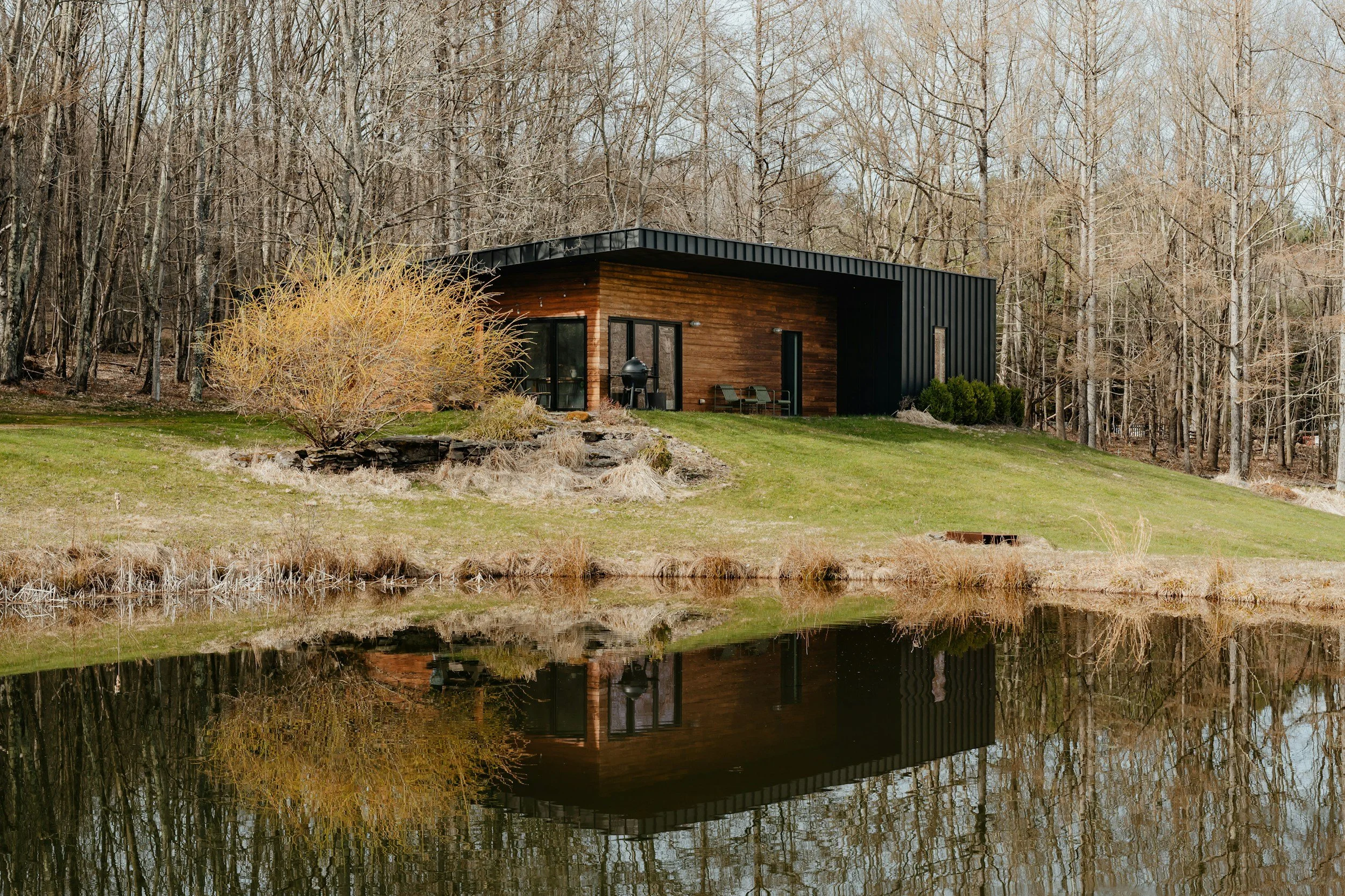 A small modern house with black metal siding and wooden accents, situated next to a pond with its reflection visible in the water, surrounded by leafless trees in a natural setting.