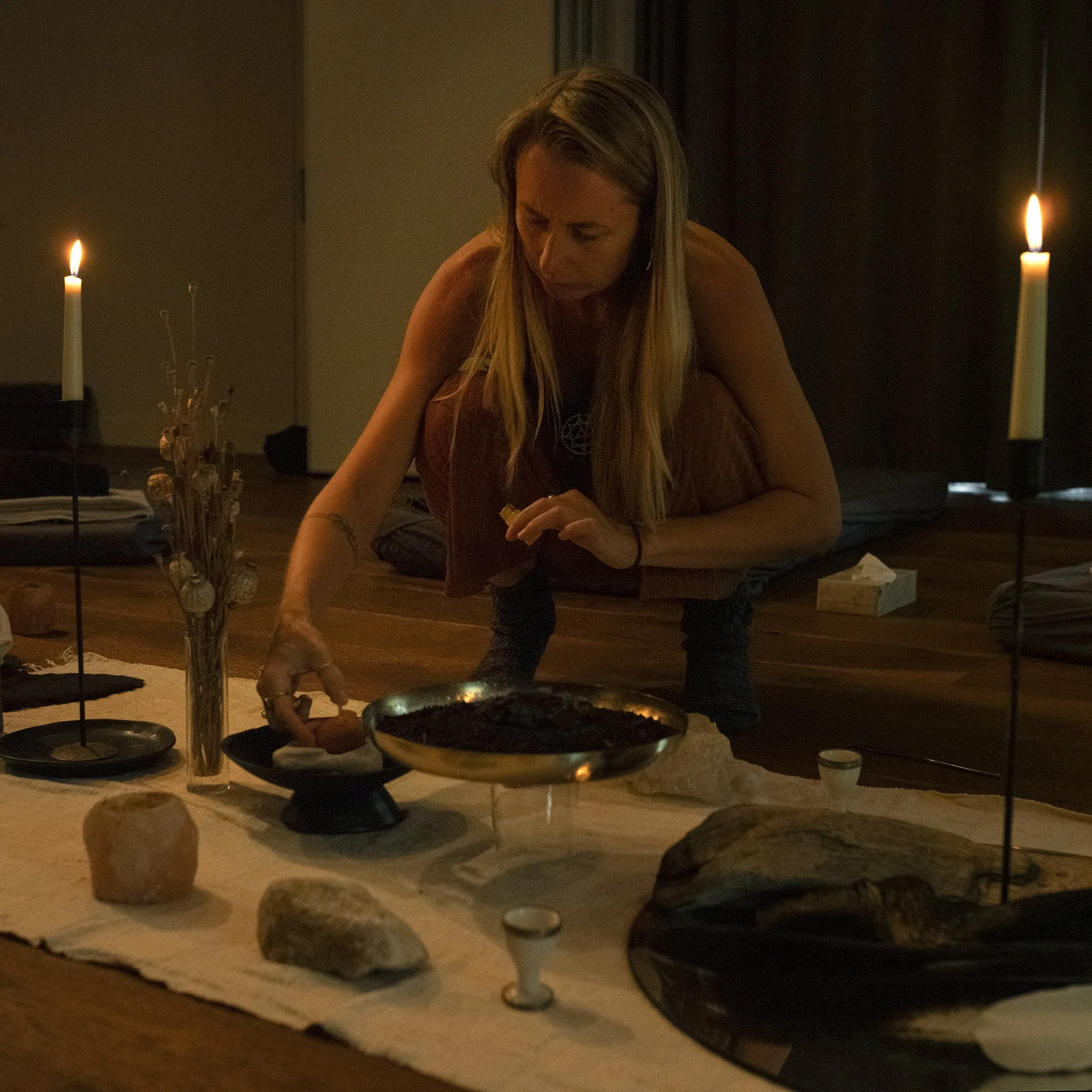 A woman with long blonde hair sitting at a table in a dimly lit room, surrounded by candles and rocks, with a dark background.