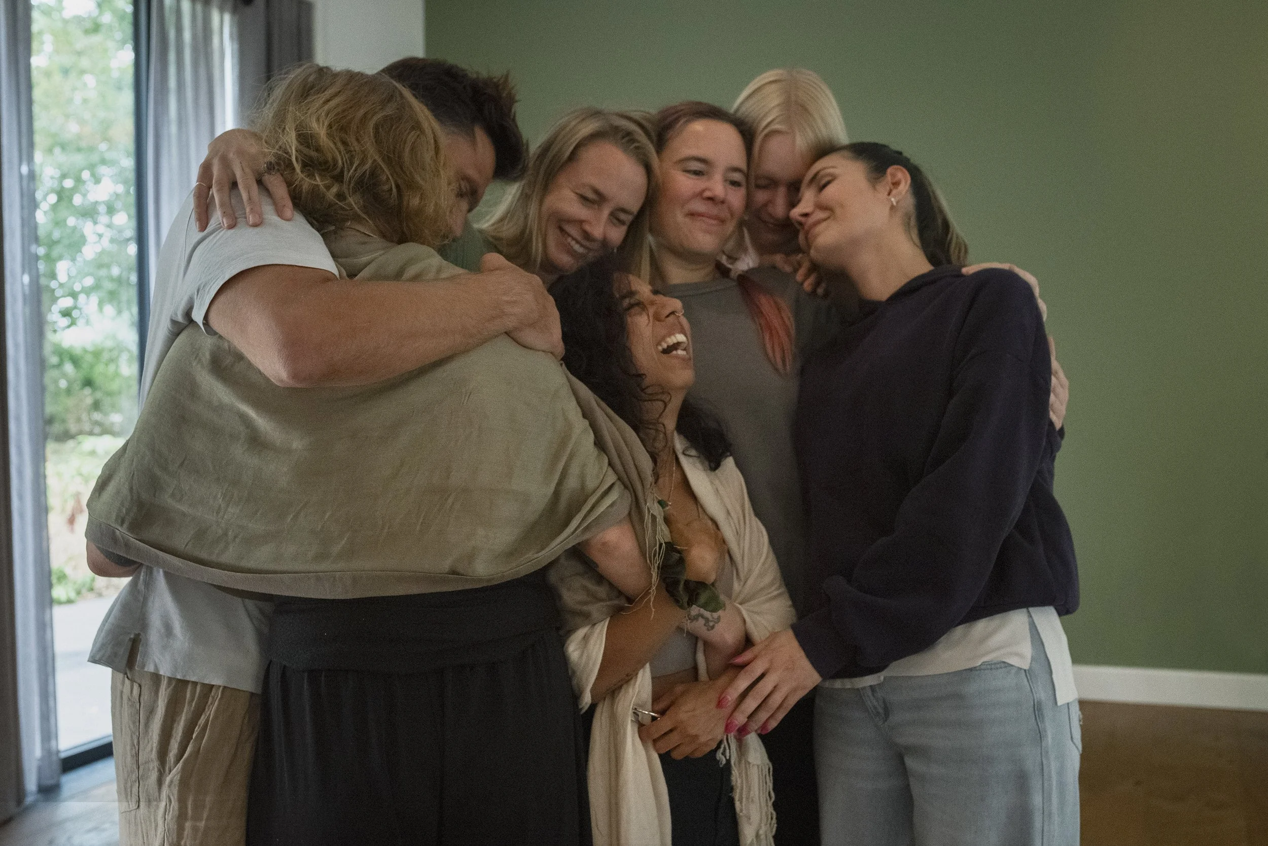Group of six diverse women embracing each other in a circle indoors, smiling and showing closeness and friendship.