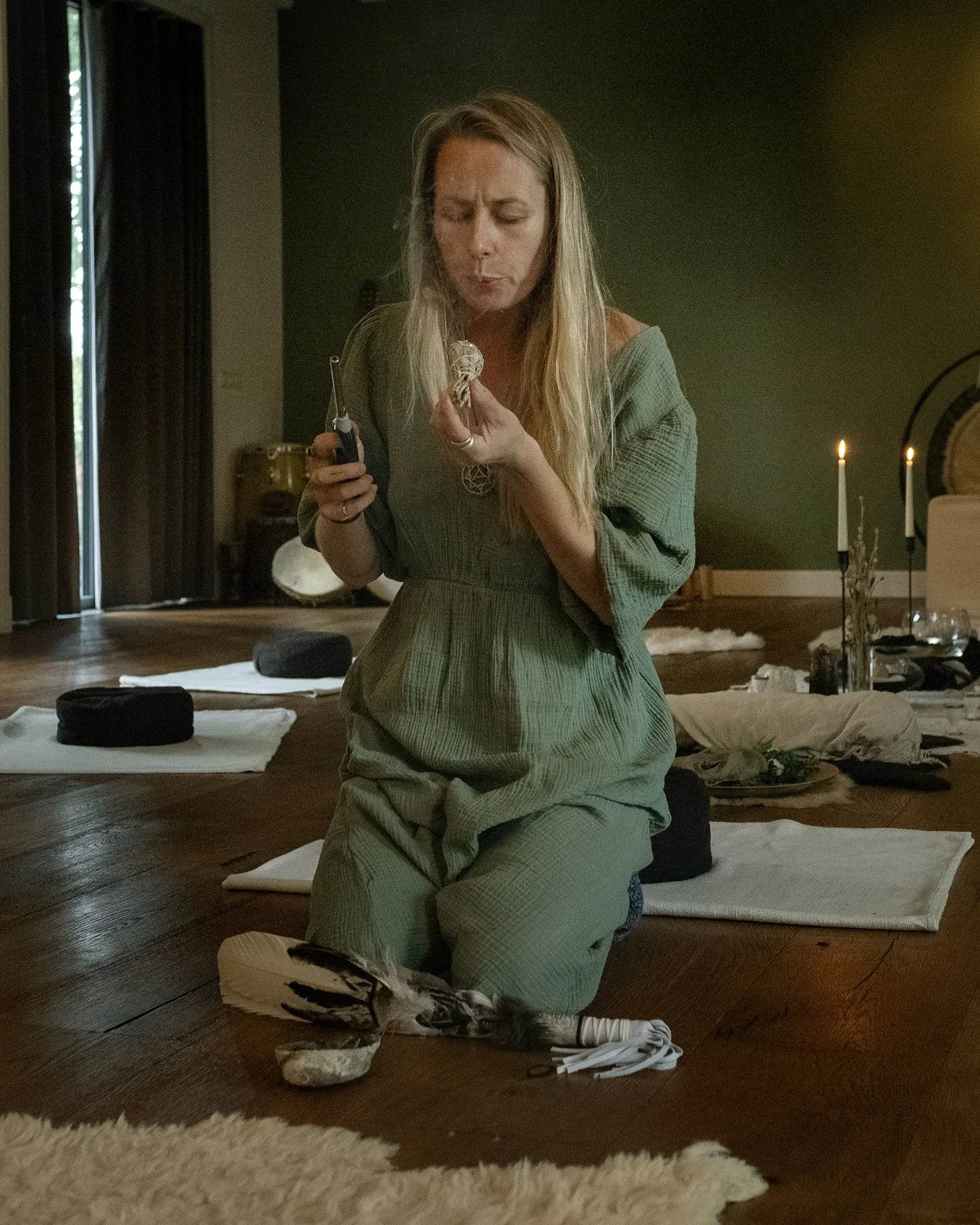 A woman kneeling on a wooden floor, holding a small object and a tool, with meditation cushions and candles in a softly lit room.