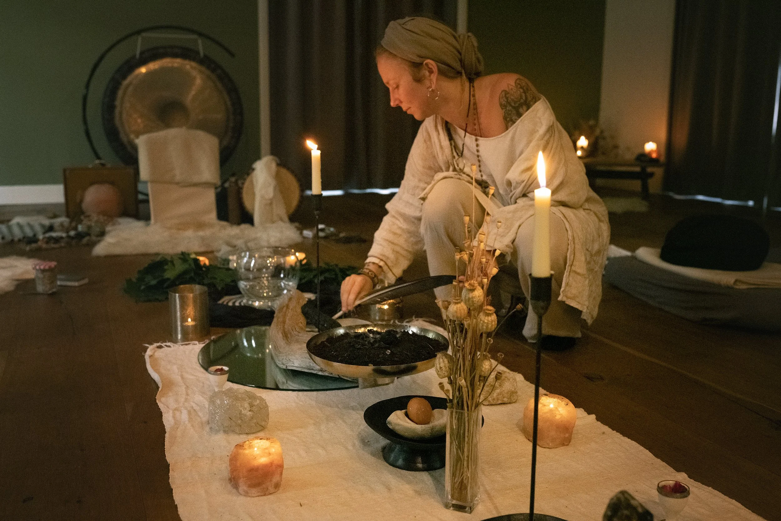 A woman with a headscarf, tattoos, and earrings is sitting at a table in a dimly lit room, surrounded by lit candles and various decorative objects, engaging in an activity.