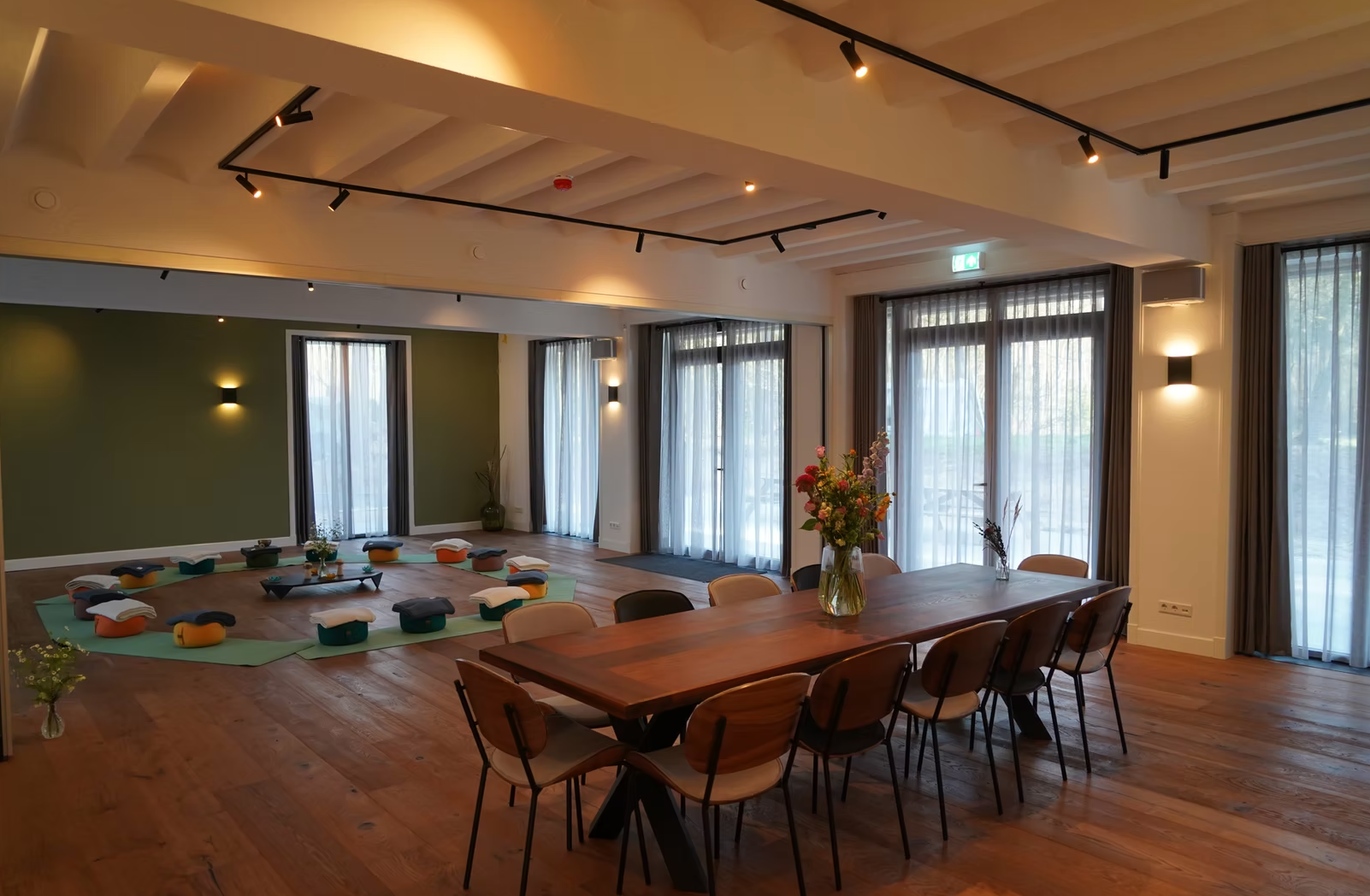 A spacious room with wooden flooring, large windows with sheer curtains, a green accent wall, and a dining table with chairs. There is a yoga or meditation area with cushions arranged in a circle on a green mat, and a vase of flowers on the table.