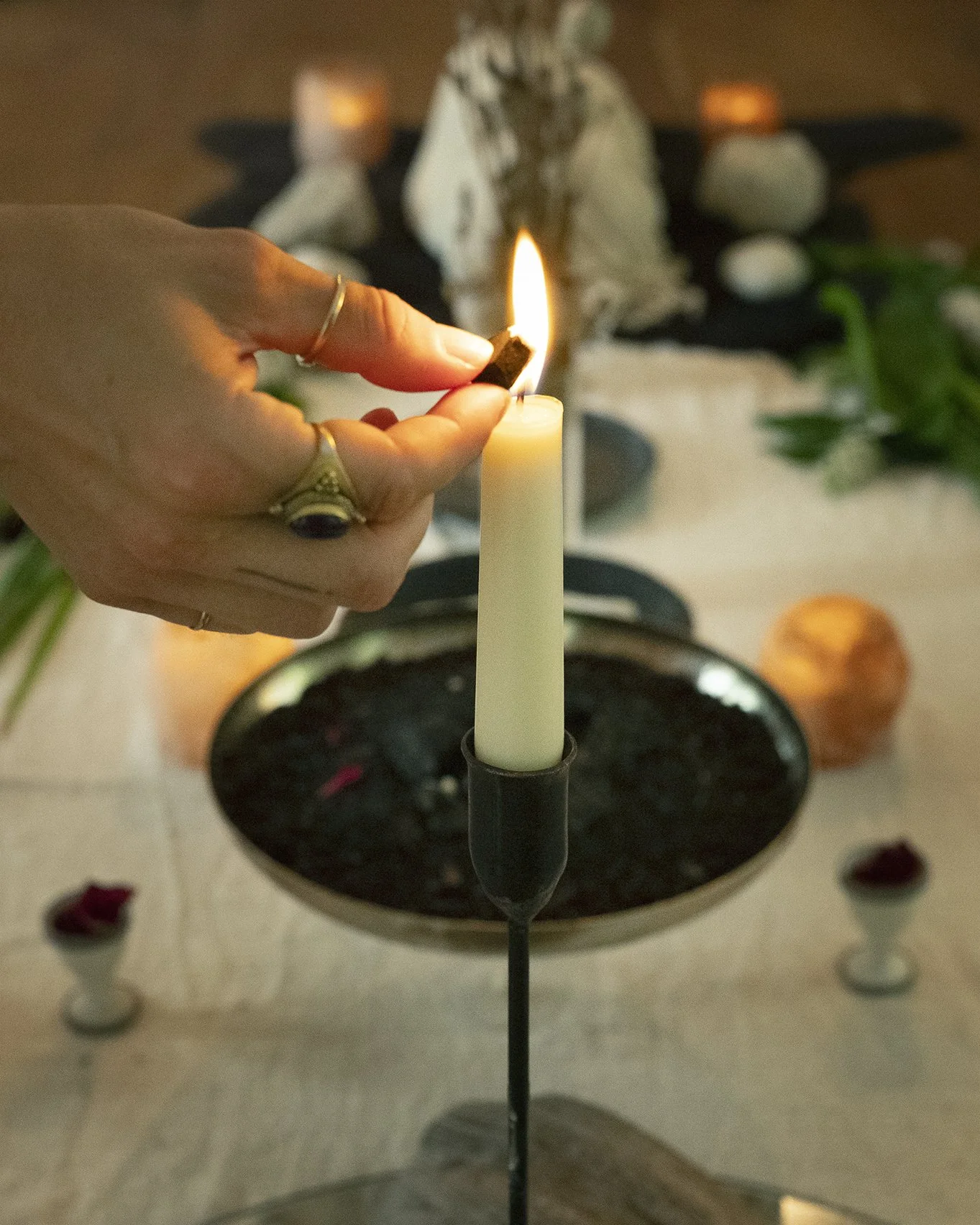 A person lighting a white candle with a match stick during a ritual or ceremony. There are two other lit candles and decorative stones on a table with a cloth, plants, and rose petals around.