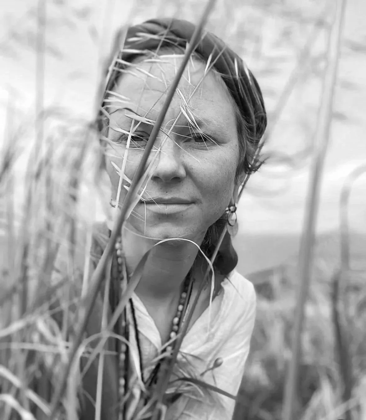Black and white photo of a woman with short hair, wearing earrings and a beaded necklace, looking through tall grass or wheat in an outdoor field.