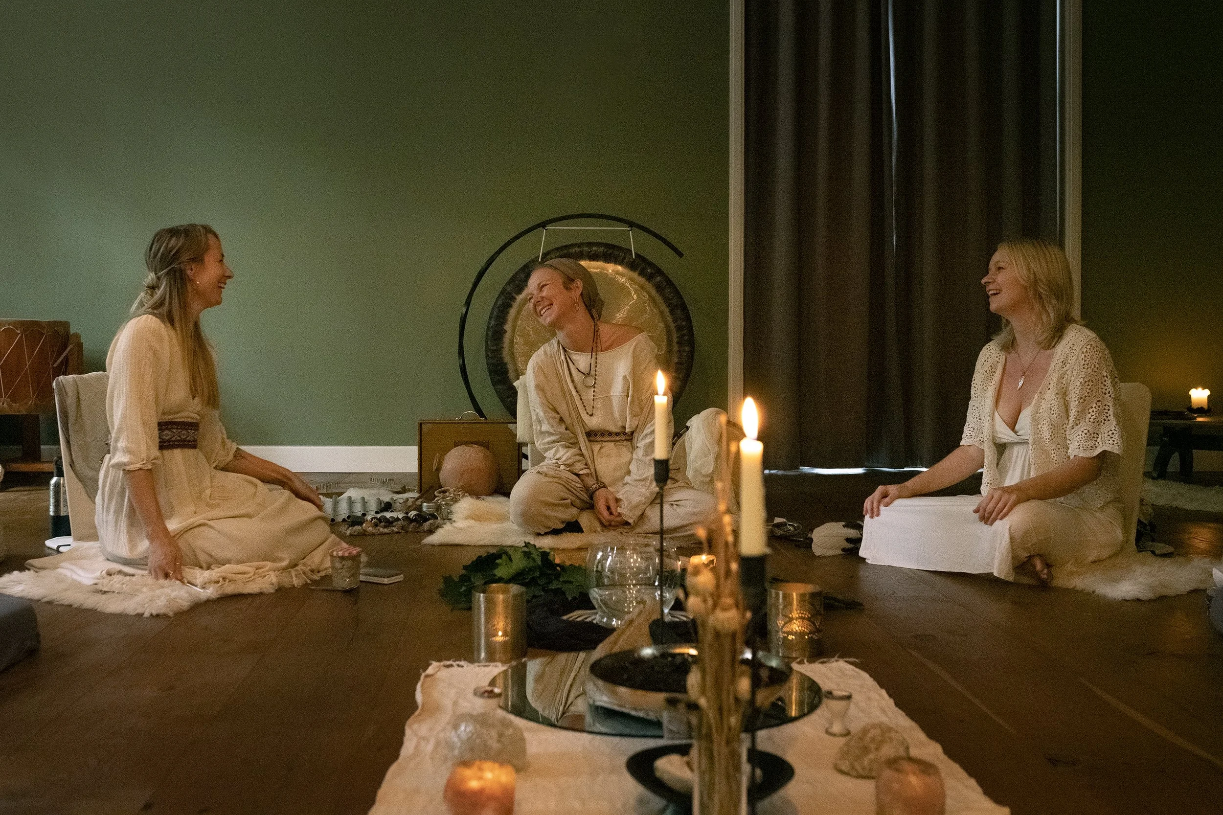 Three women sitting on the floor around a table with candles, a gong, and spiritual objects, smiling and engaging in a bonding activity in a cozy room with candles and a green wall.