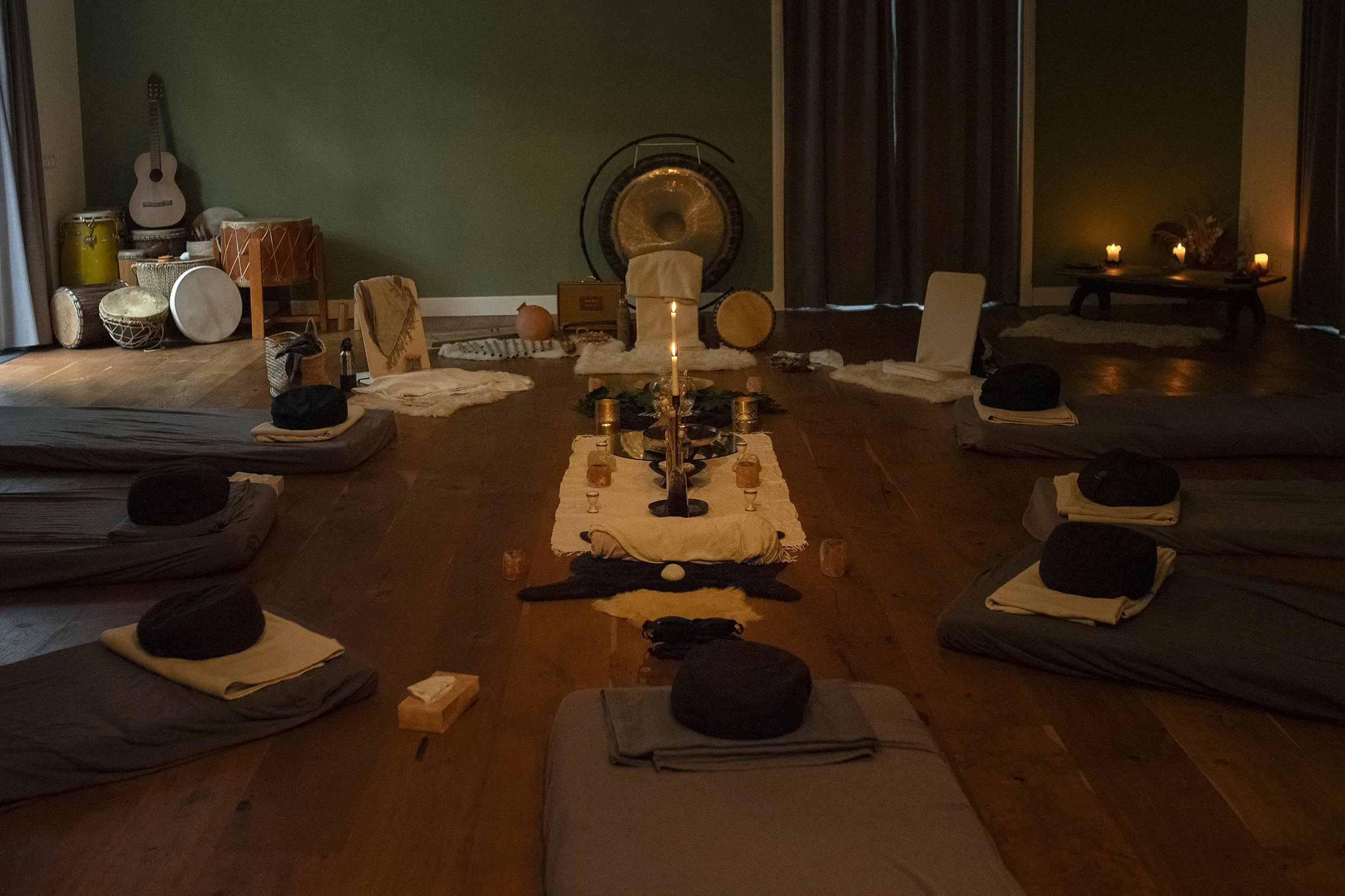 A dimly lit room set up for a ritual or meditation with cushions around a central table decorated with candles, greenery, and stones, with musical instruments and a gong in the background.