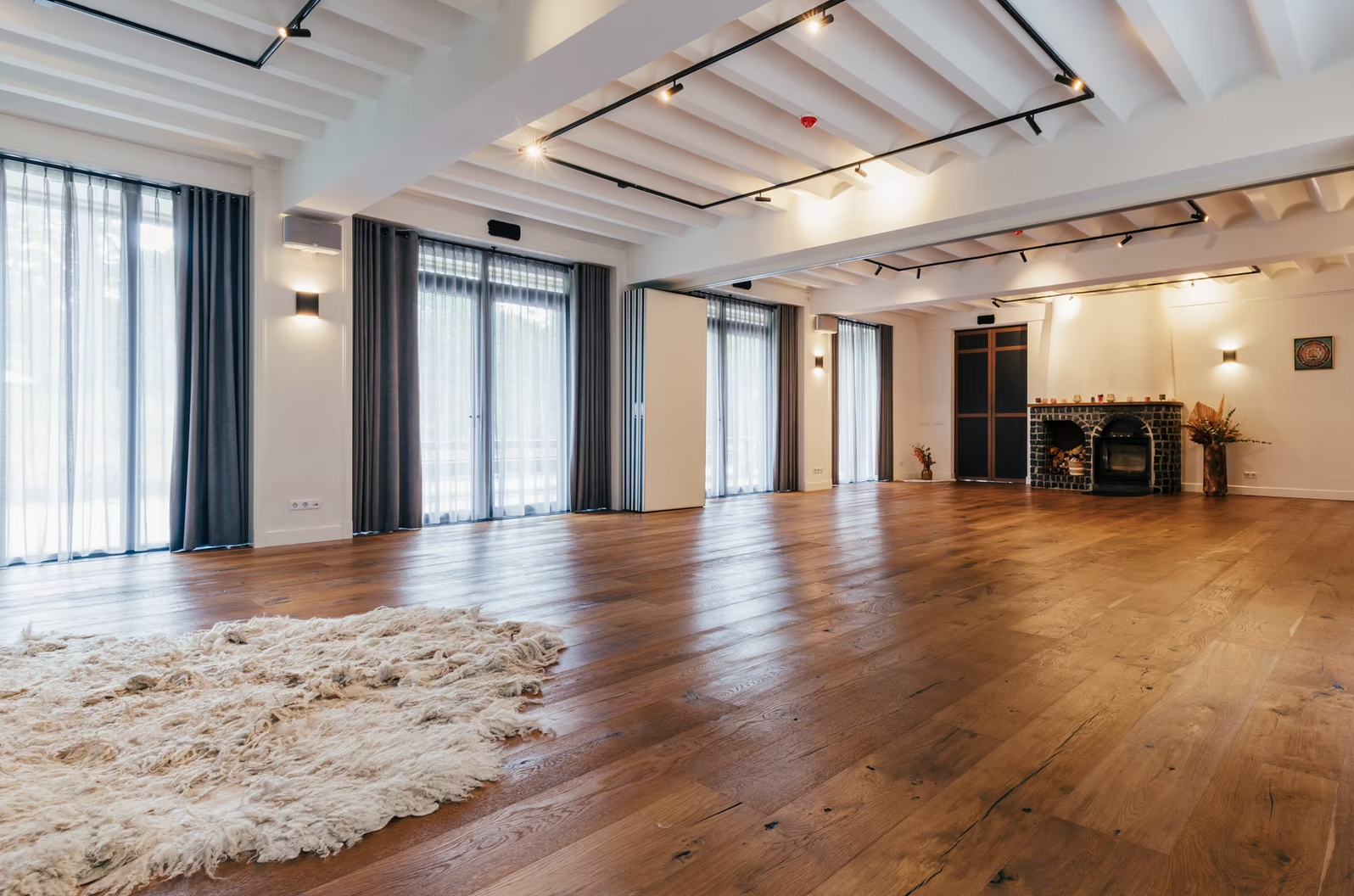 Spacious empty living room with hardwood floors, large windows with sheer curtains, a fireplace, and decorative items.