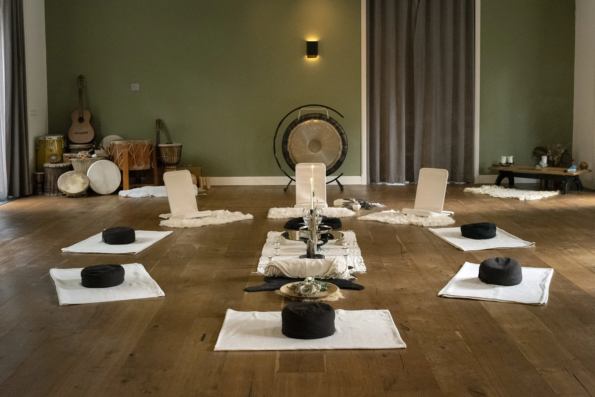 A meditation room with a green wall, helping to create a calming atmosphere. The room displays musical instruments like guitars, drums, and a gong, possibly for musical therapy or relaxation sessions. A low table on the right side has candles and decorative items, enhancing the serene environment. The center of the room has neatly arranged white cushions with black round bolsters on each, placed on small white mats, indicating a space designated for seated meditation or yoga practice. The warm lighting from a wall sconce adds to the peaceful ambiance.