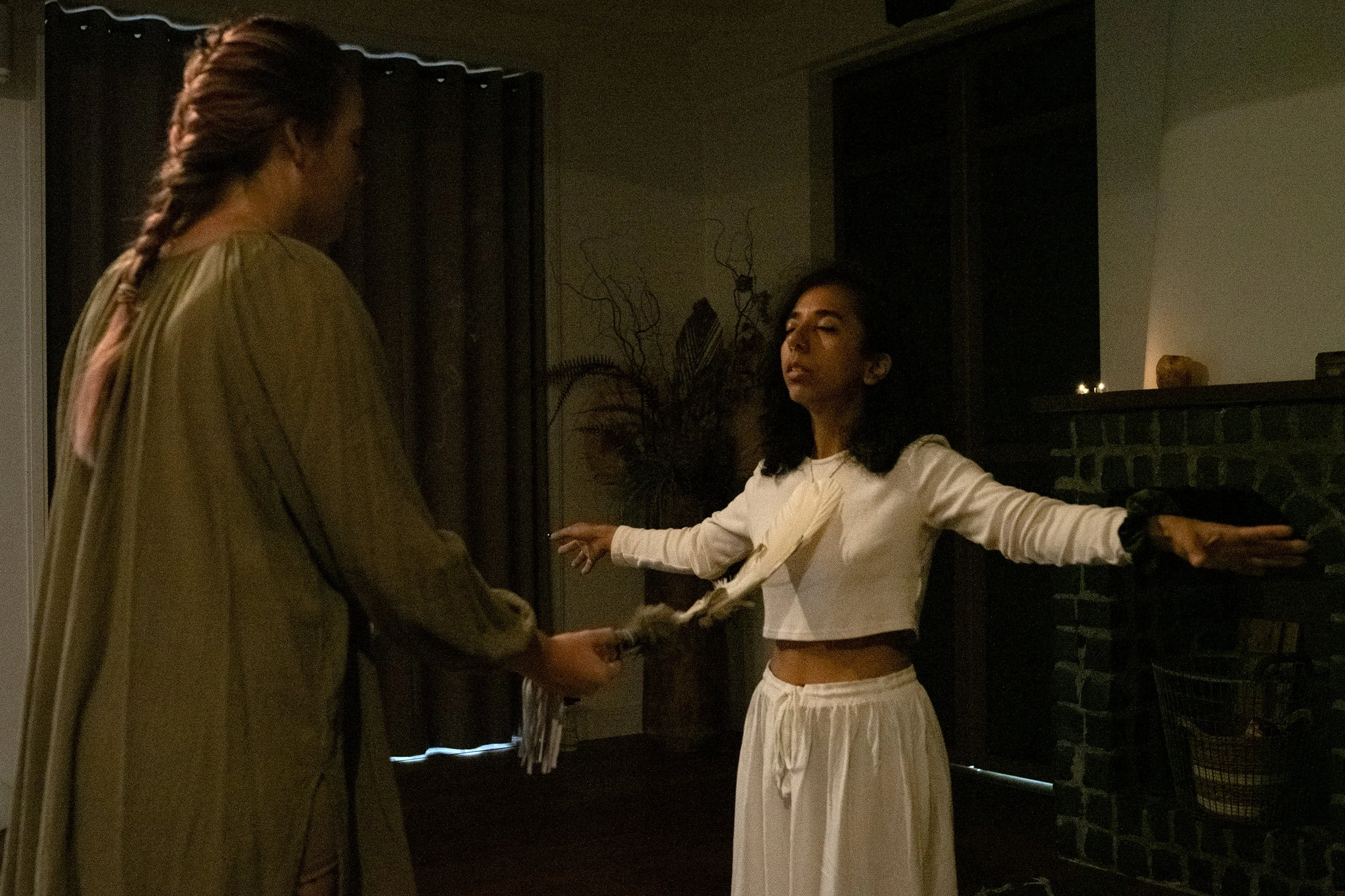 Two women in a dimly lit room, one standing and holding papers, the other sitting with arms outstretched, behind a dark fireplace with a candle and a decorative stone on the mantel.