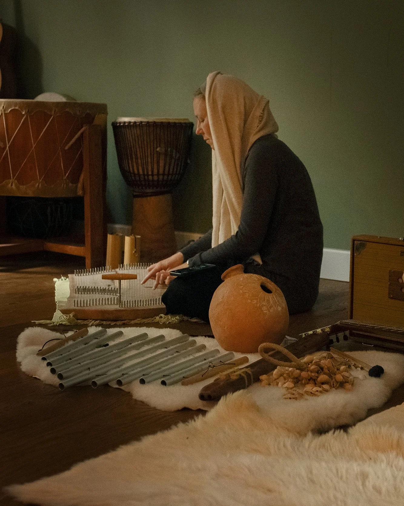 A woman wearing a headscarf is sitting on the floor, playing a musical instrument with metal tines. In front of her are various musical instruments and objects, including a clay pot, a set of pipes, and small percussion instruments, on a white furry rug.